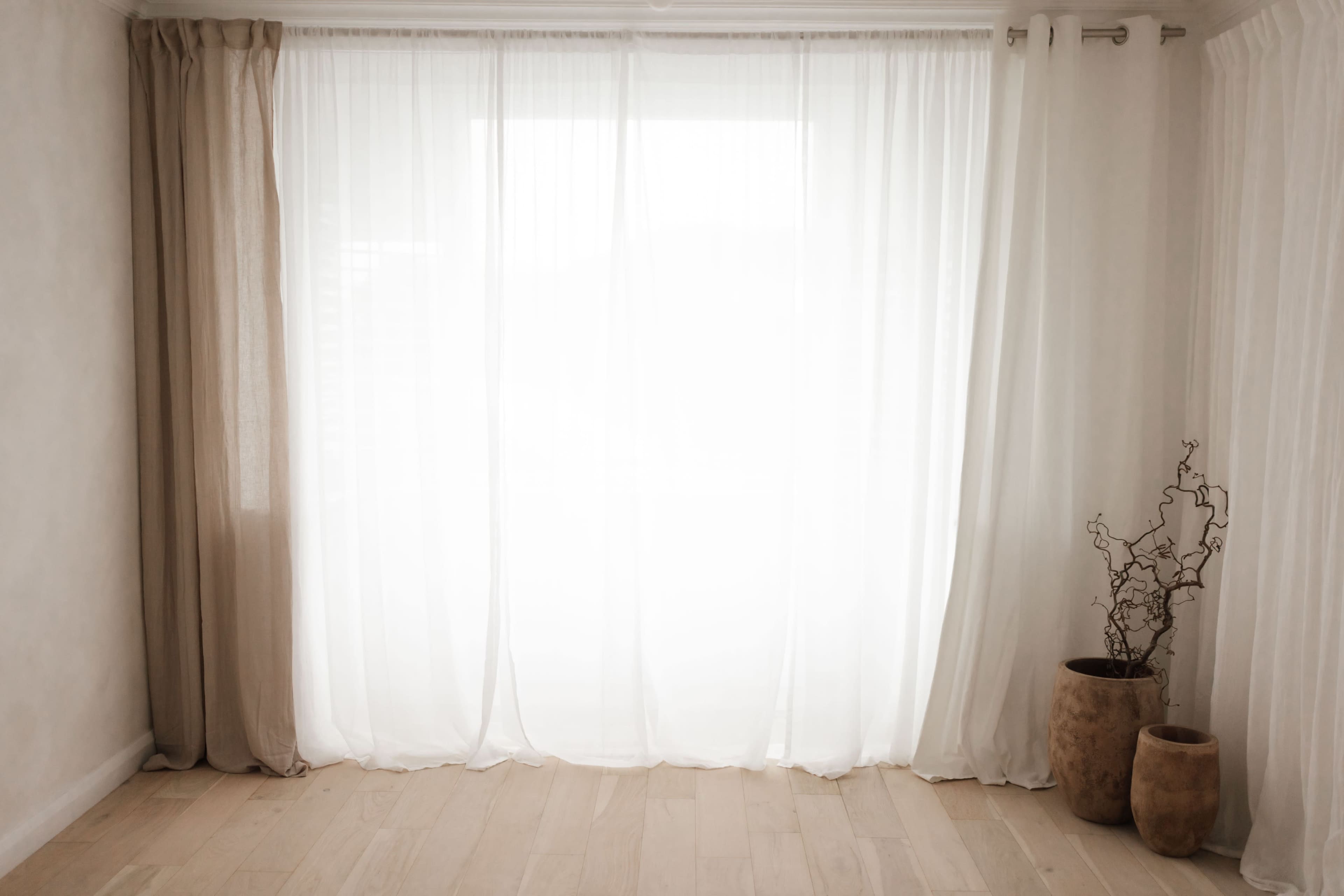 The image shows a softly lit room with sheer white curtains, wooden flooring, and two decorative pots, one containing a branch.