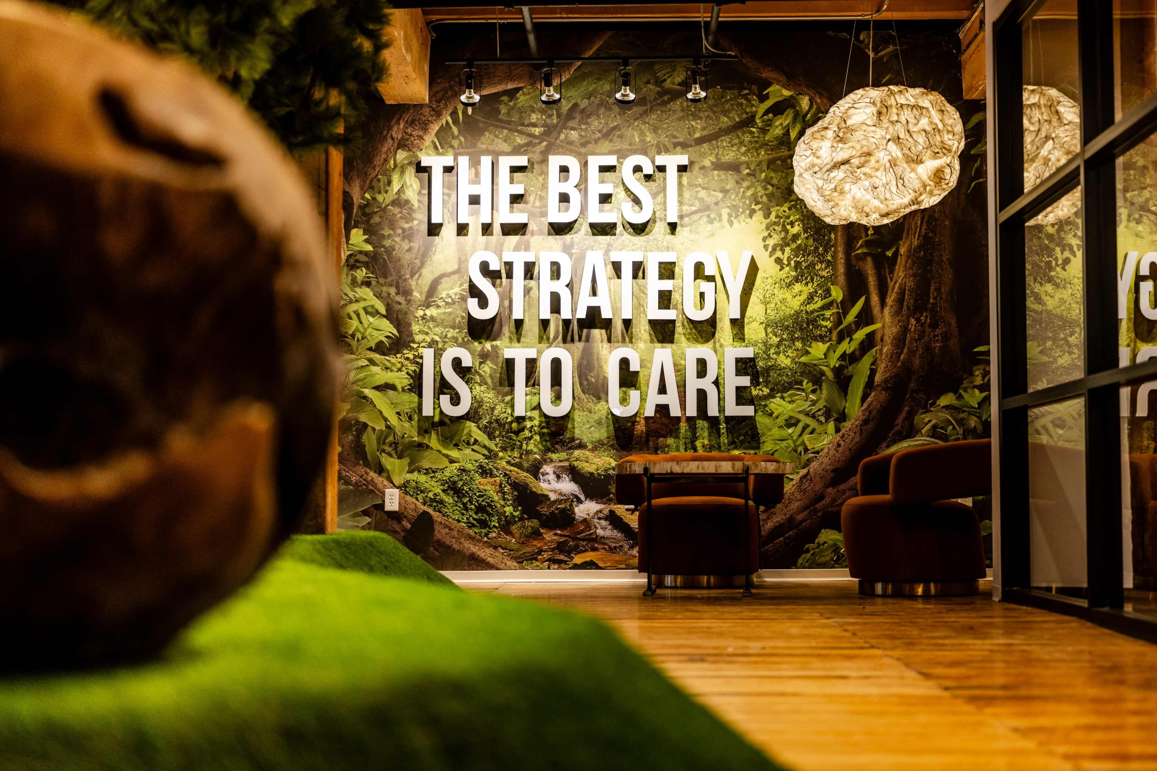 The image features a modern interior space with a large motivational quote on the wall, surrounded by greenery and a wooden floor.