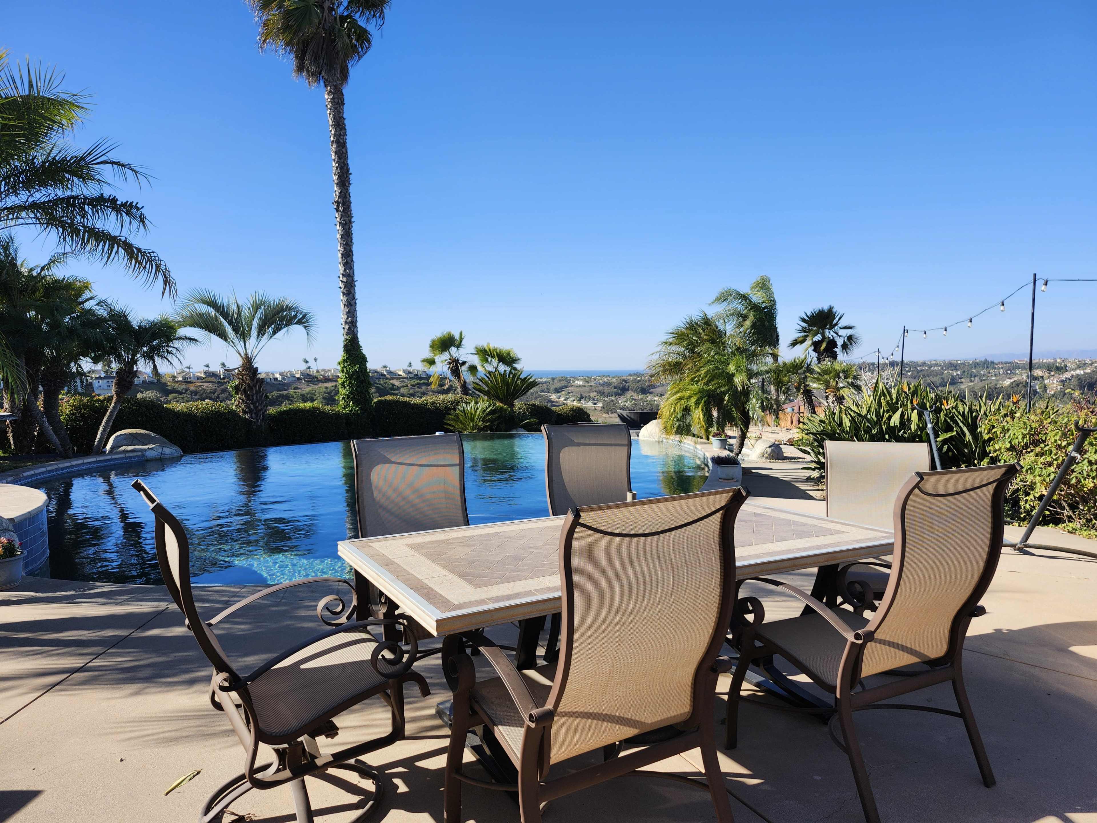 A patio table with chairs overlooks a swimming pool and a view of the landscape under a clear blue sky.