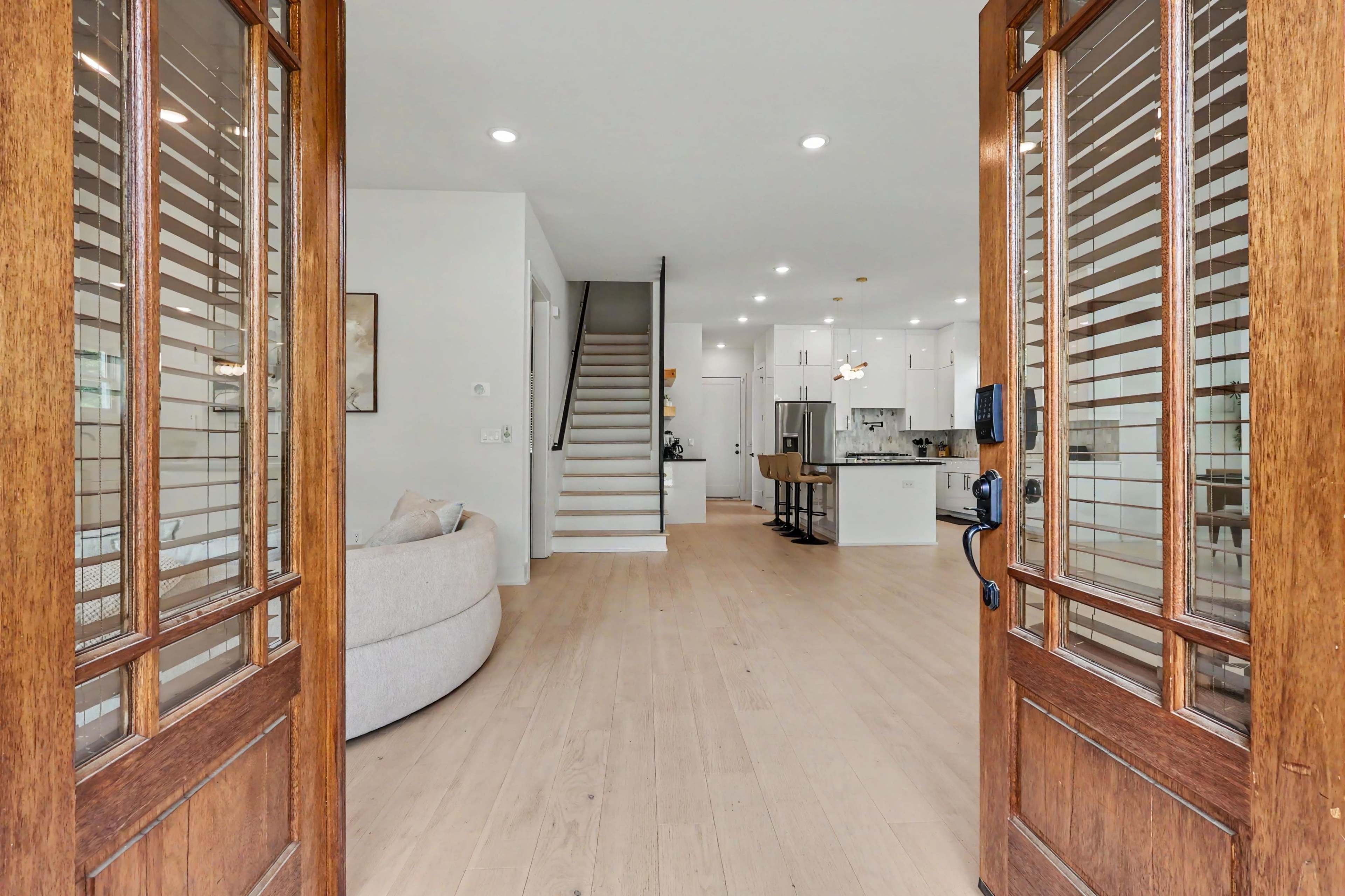 The image shows an open entrance with wooden double doors leading into a bright, modern living space featuring a staircase and a kitchen area.