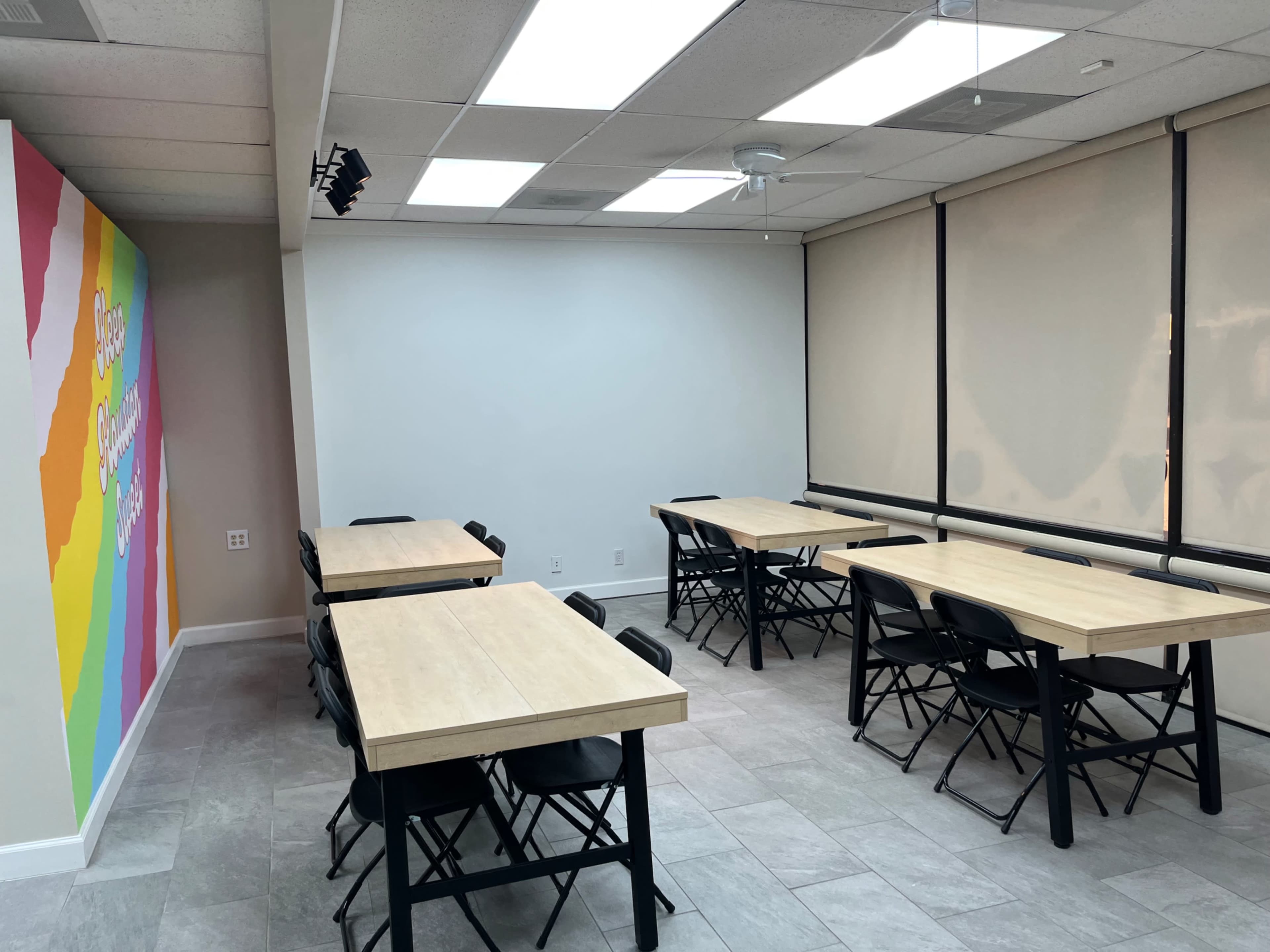 The image shows a bright room with several wooden tables and black folding chairs arranged in a classroom-style layout, accompanied by a colorful mural on one wall.