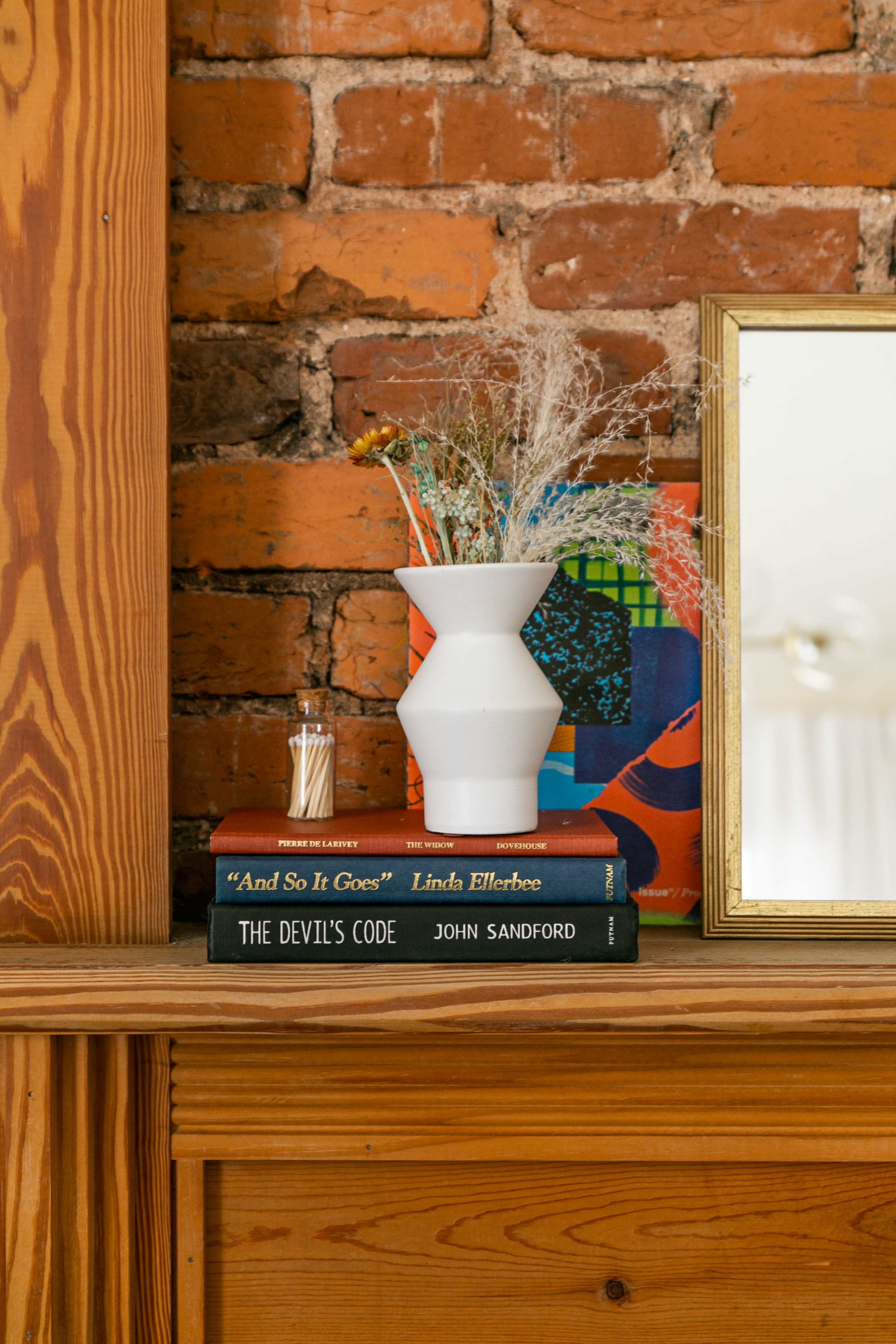 A white vase filled with dried flowers sits on a stack of books, next to a small mirror, against a backdrop of exposed brick.