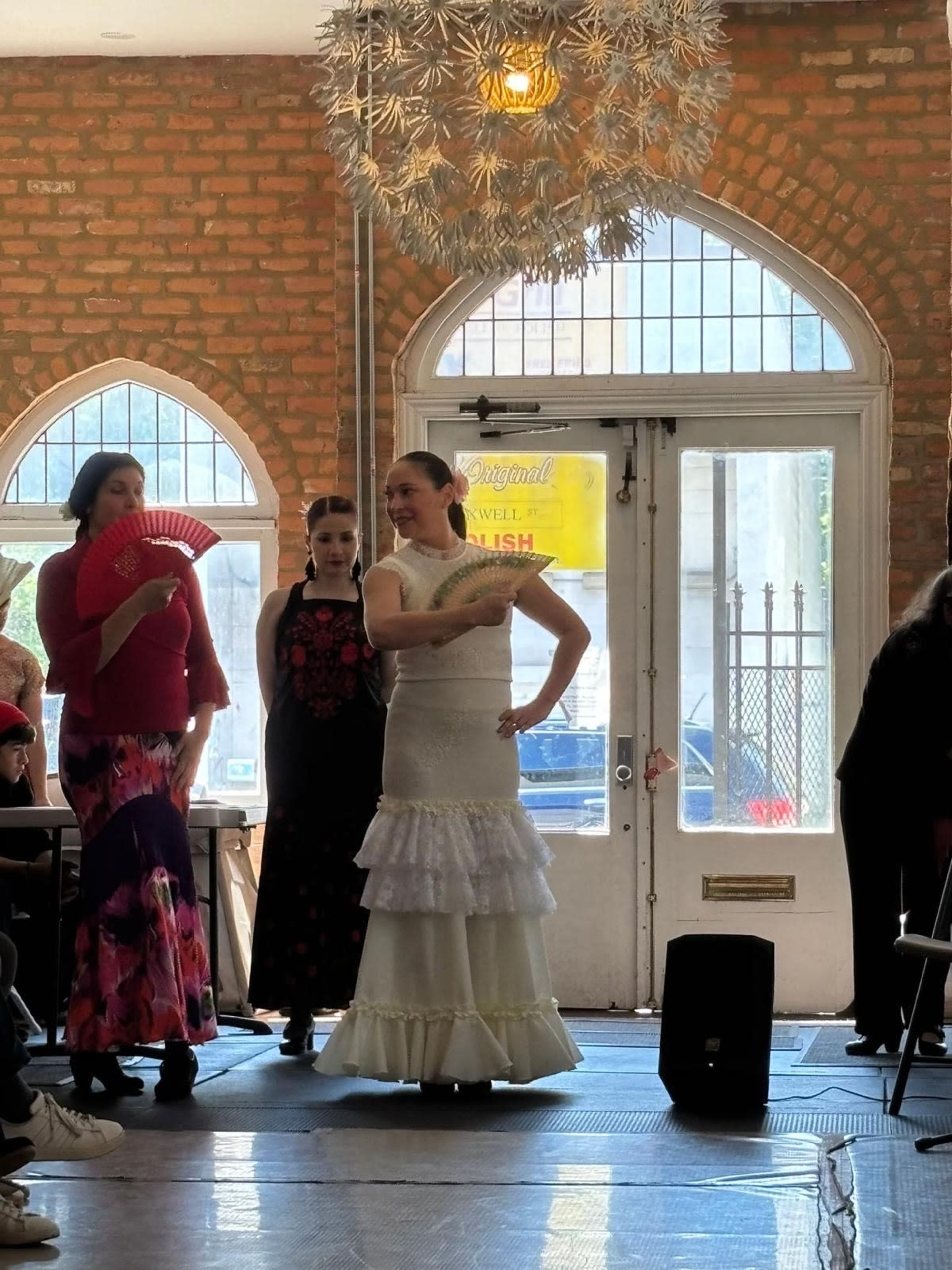 A woman in a white dress performs flamenco dance in front of an audience, with two other women nearby, one holding a fan and the other wearing a black dress.
