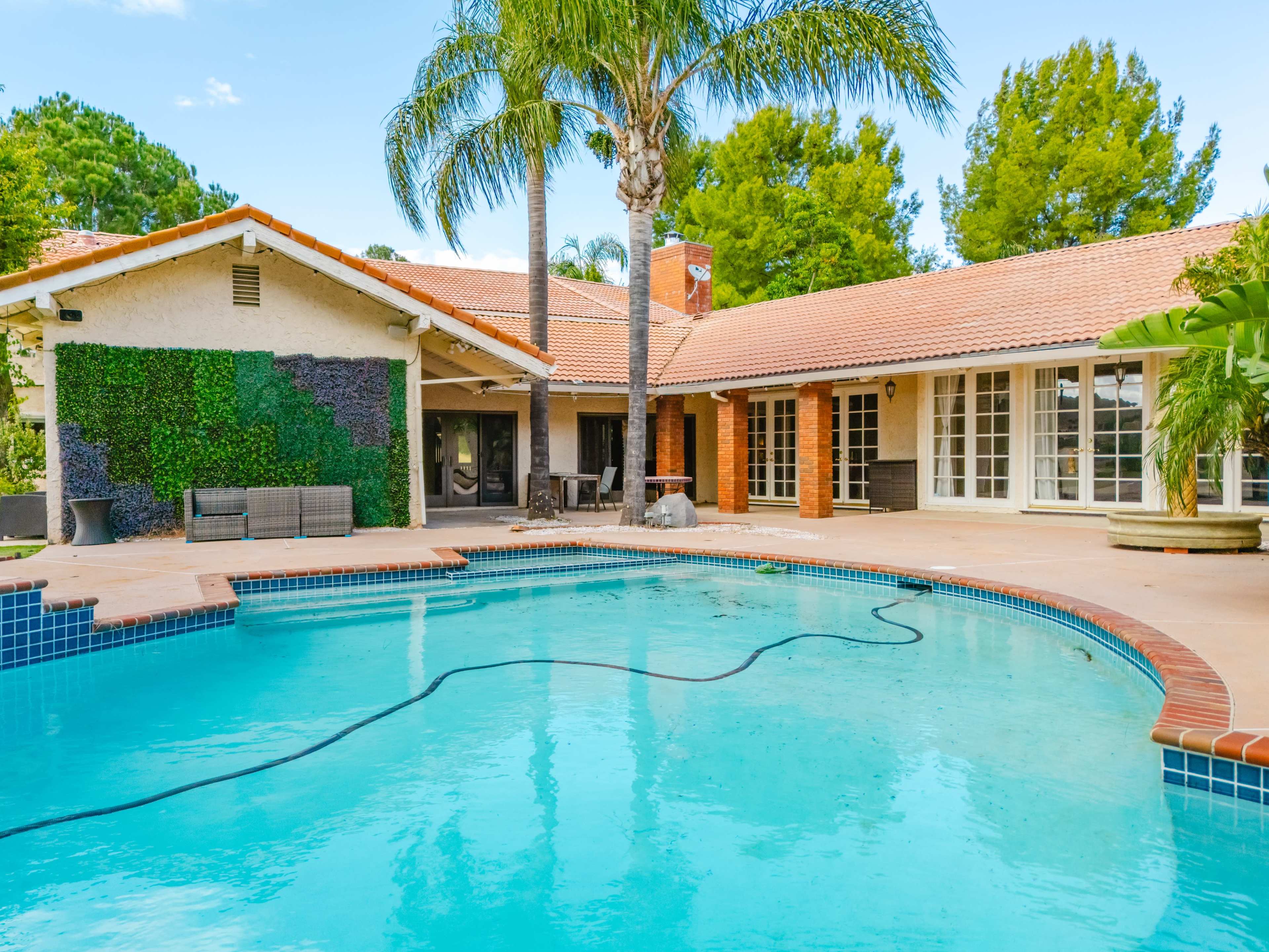 A single-story house with a tiled roof is situated beside a blue swimming pool, surrounded by palm trees and a patio area.