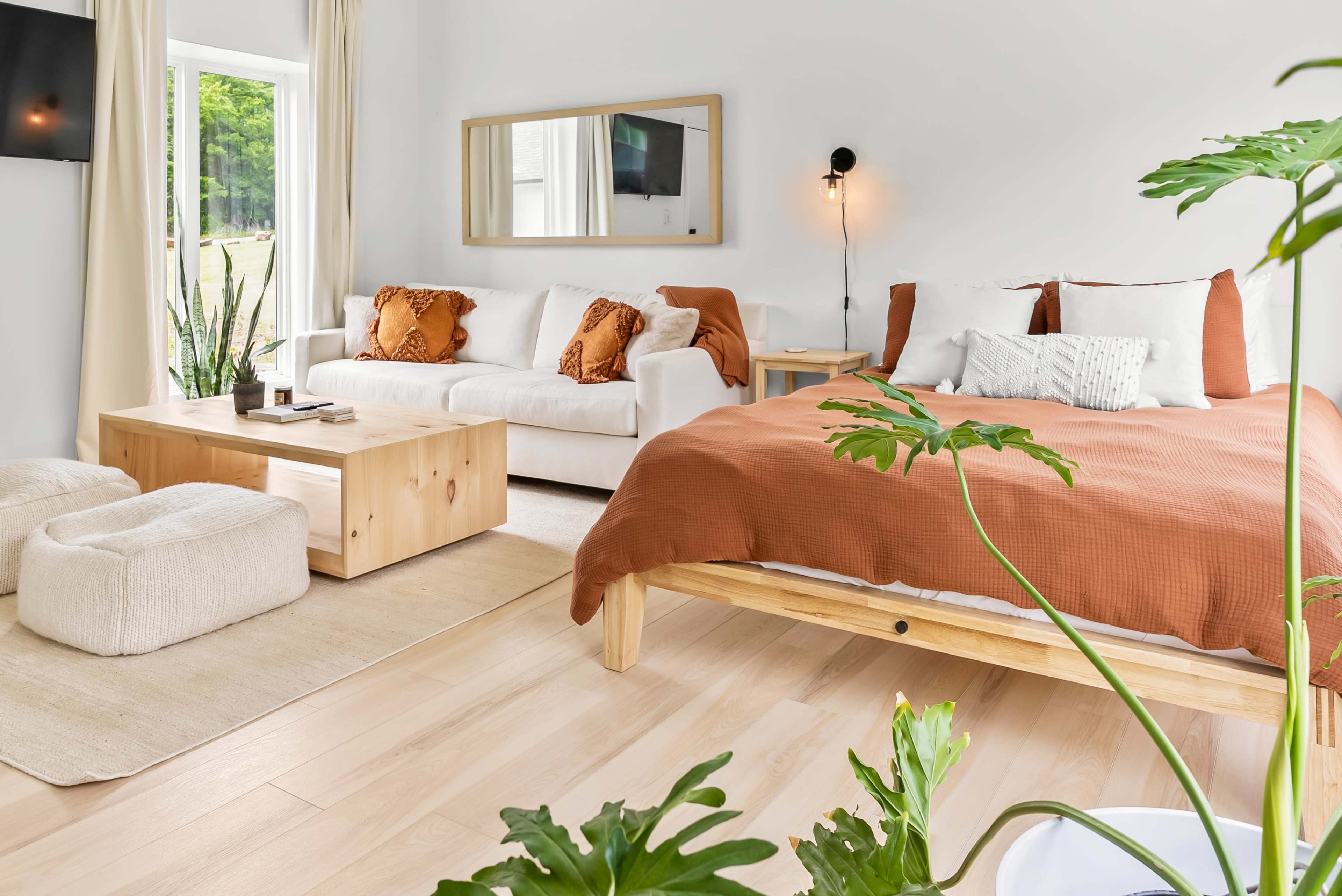 The image depicts a minimalist, bright living space featuring a bed with an orange coverlet, a white sofa, a wooden coffee table, and large windows with natural light.
