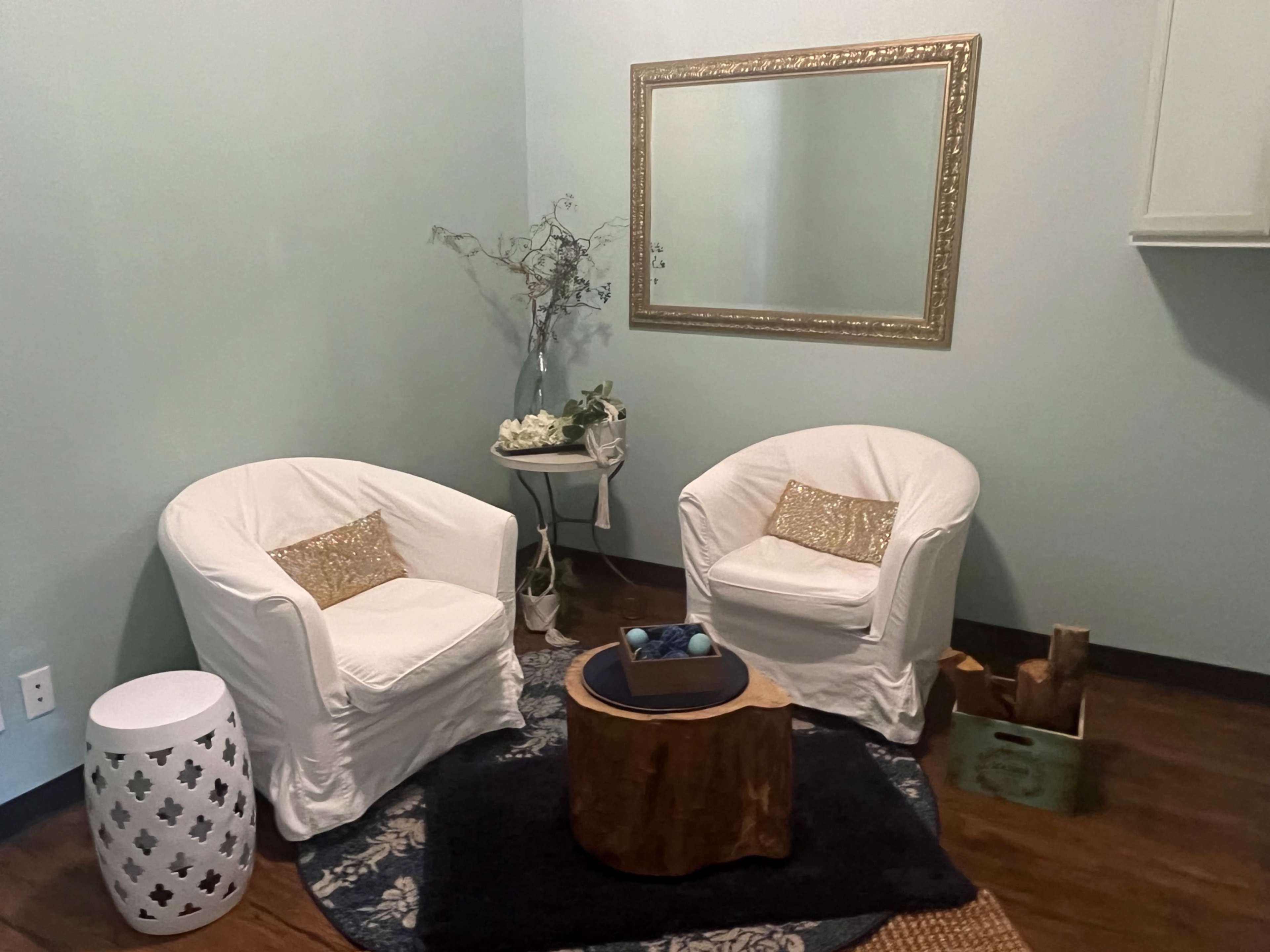 The image shows a small sitting area with two white chairs, a wooden coffee table, a dark rug, and a decorative mirror on a light green wall.