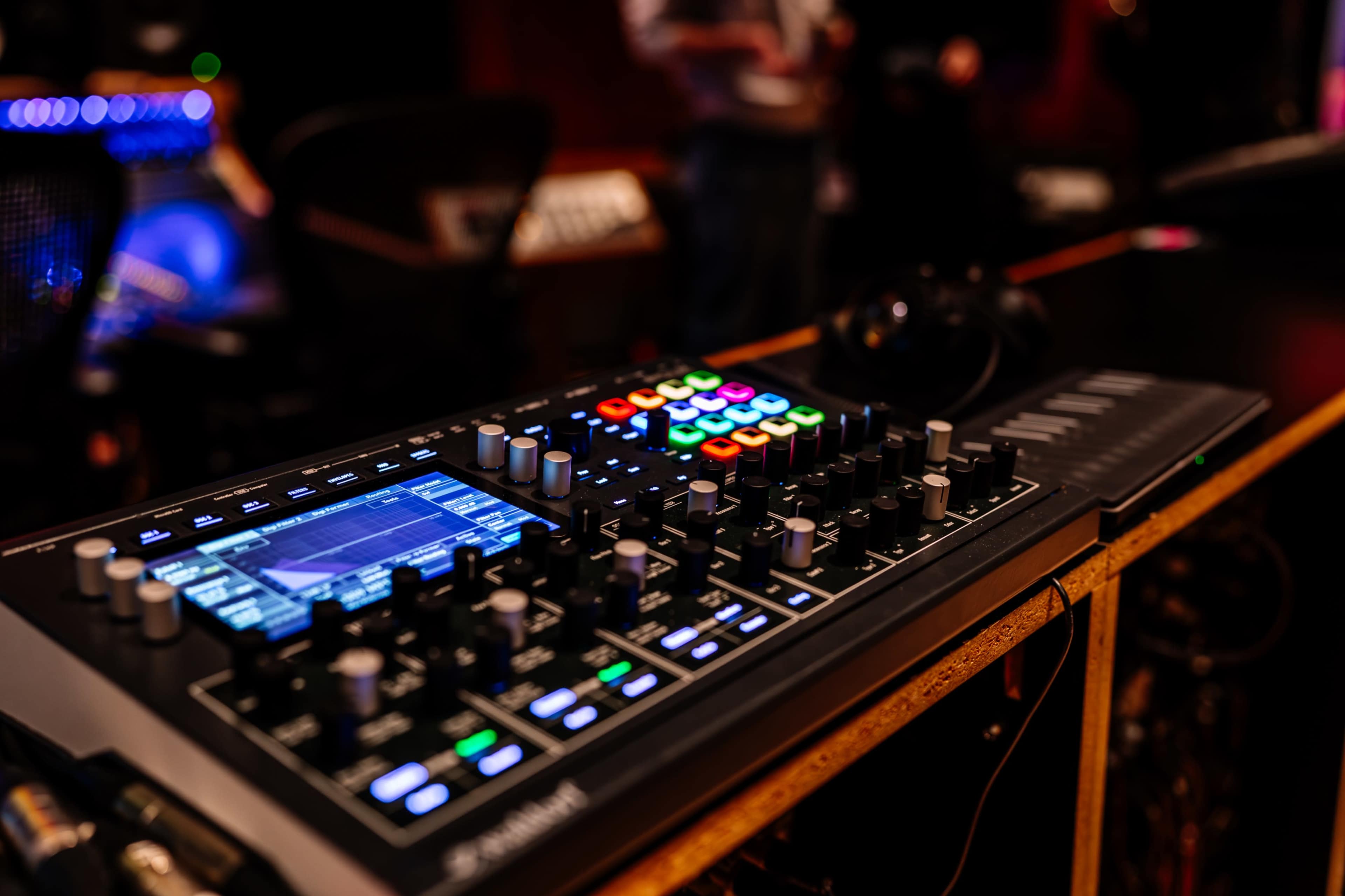 A digital audio workstation console featuring various knobs, buttons, and a visual display is positioned on a desk in a dimly lit studio.