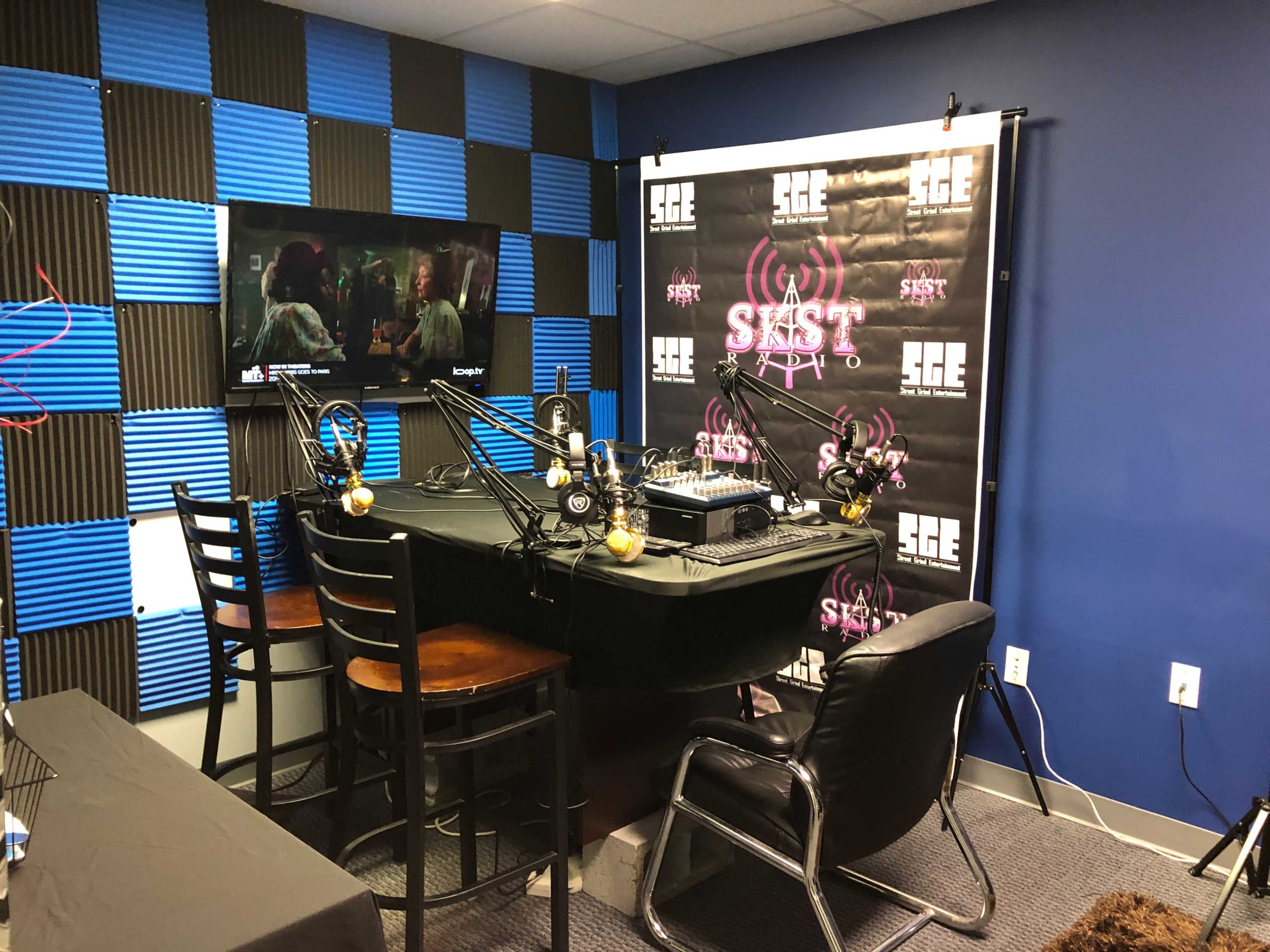 A recording studio with a black table, microphones, and chairs arranged around it, along with a television displaying a scene on the wall behind a backdrop featuring a logo.