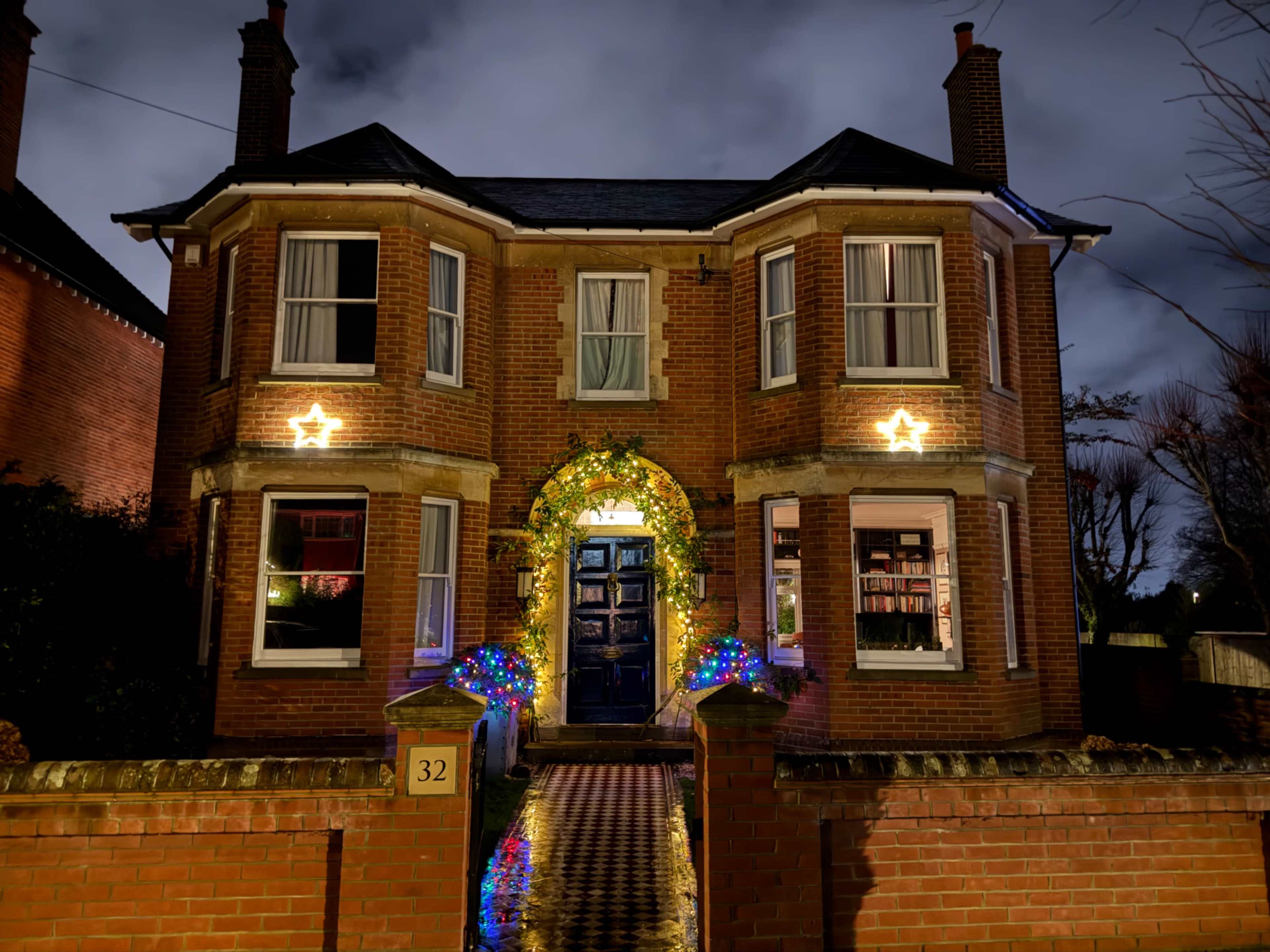 A two-story brick house, illuminated with festive decorations, features a blue front door and large windows, with lights and a wreath adorning the entrance.