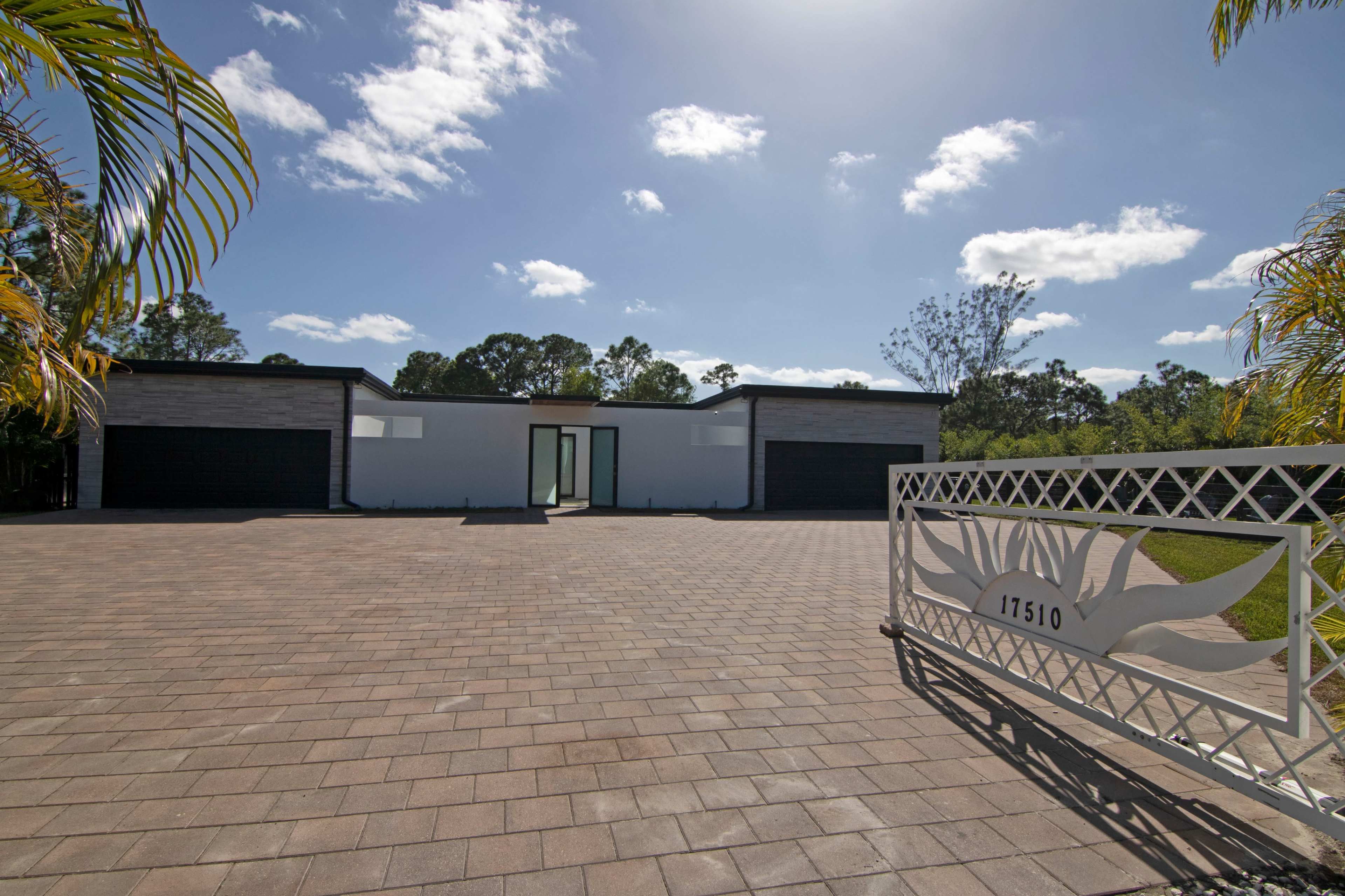 A modern house with a large paved driveway is set behind a decorative white gate and surrounded by sparse landscaping.