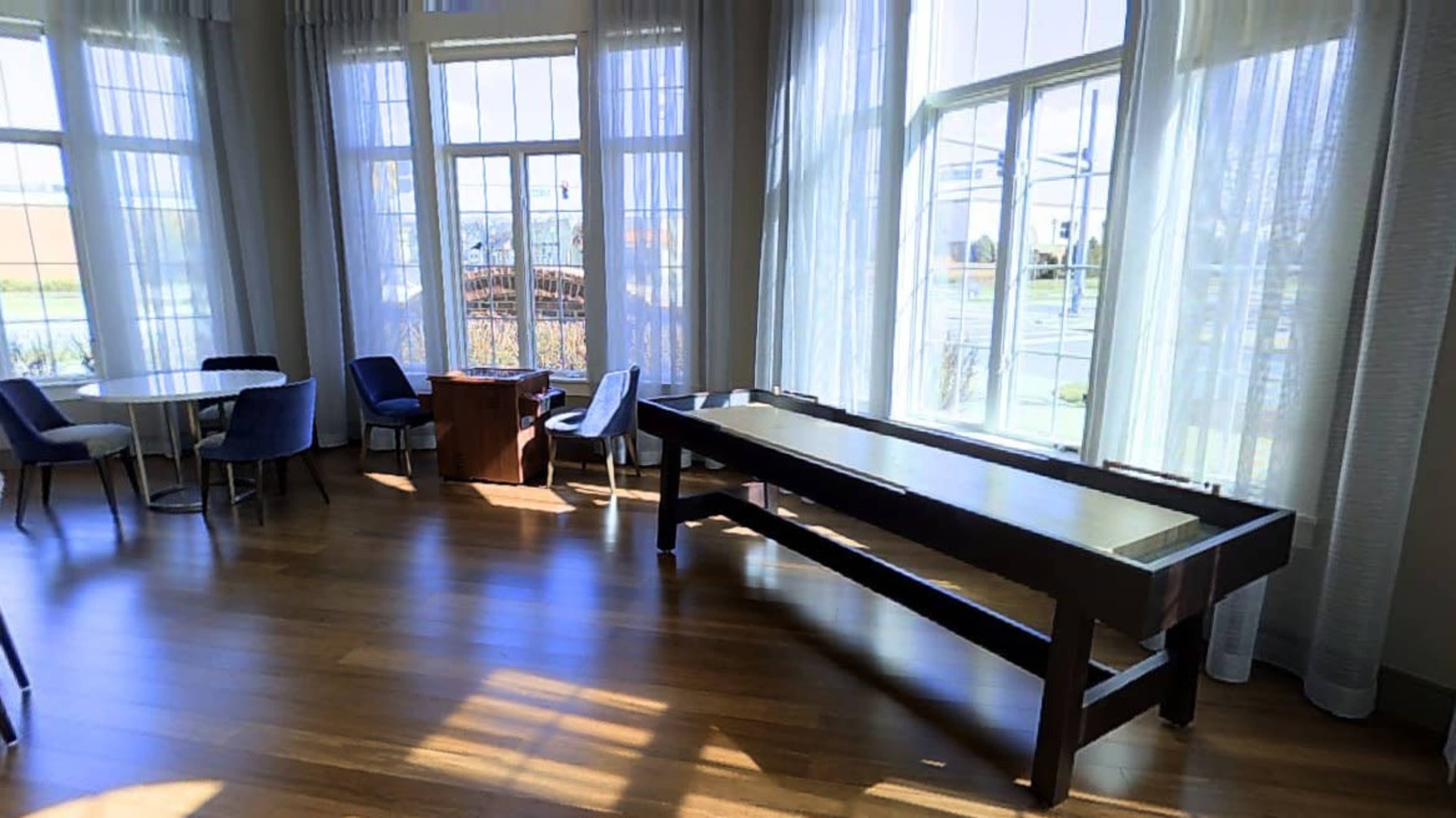 A spacious room with large windows, wooden flooring, and furniture including a table, chairs, and a shuffleboard table.