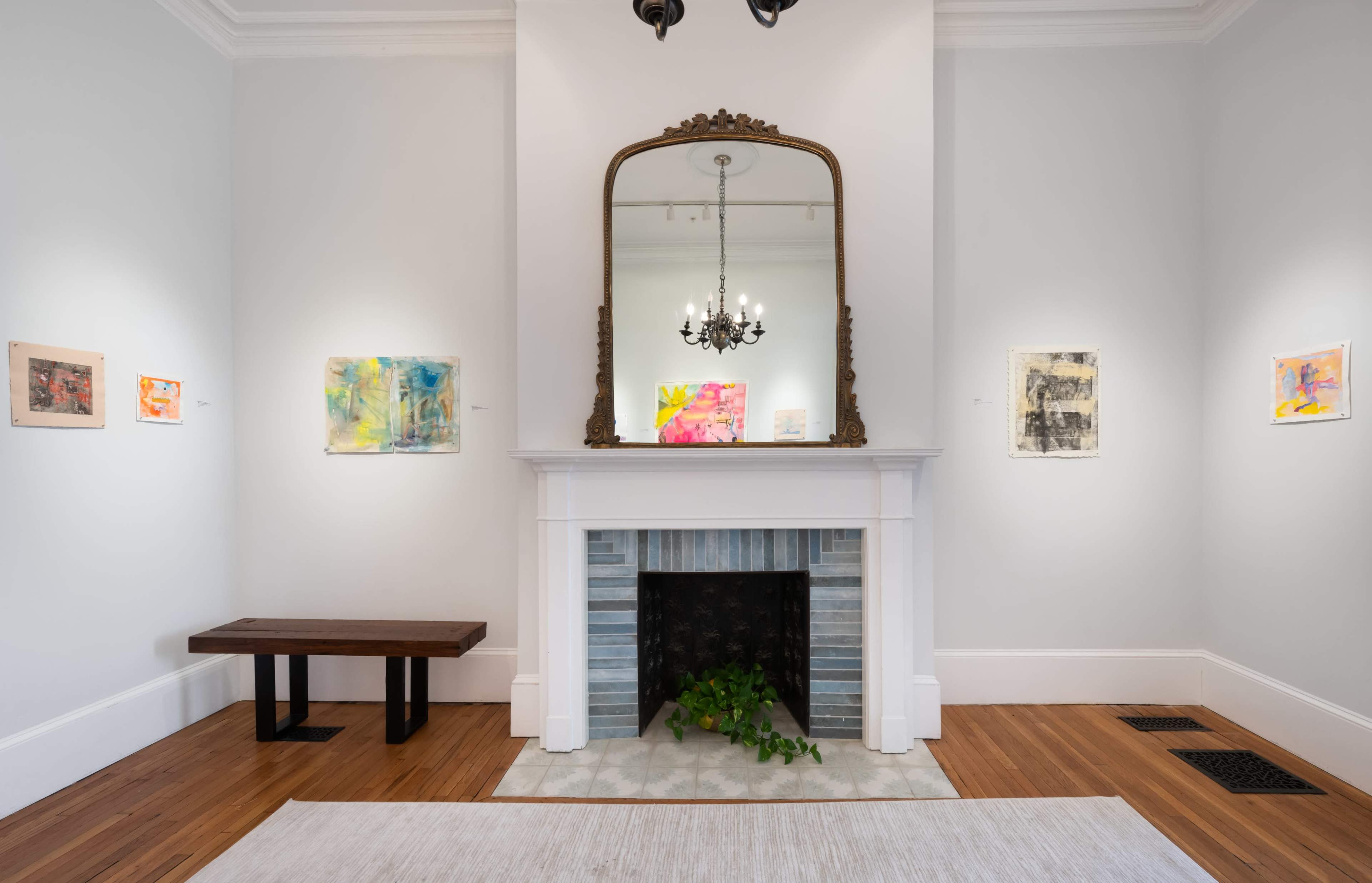 The image shows a gallery space featuring colorful abstract paintings on the walls, a mirror above a decorative fireplace, and a wooden bench in the center.
