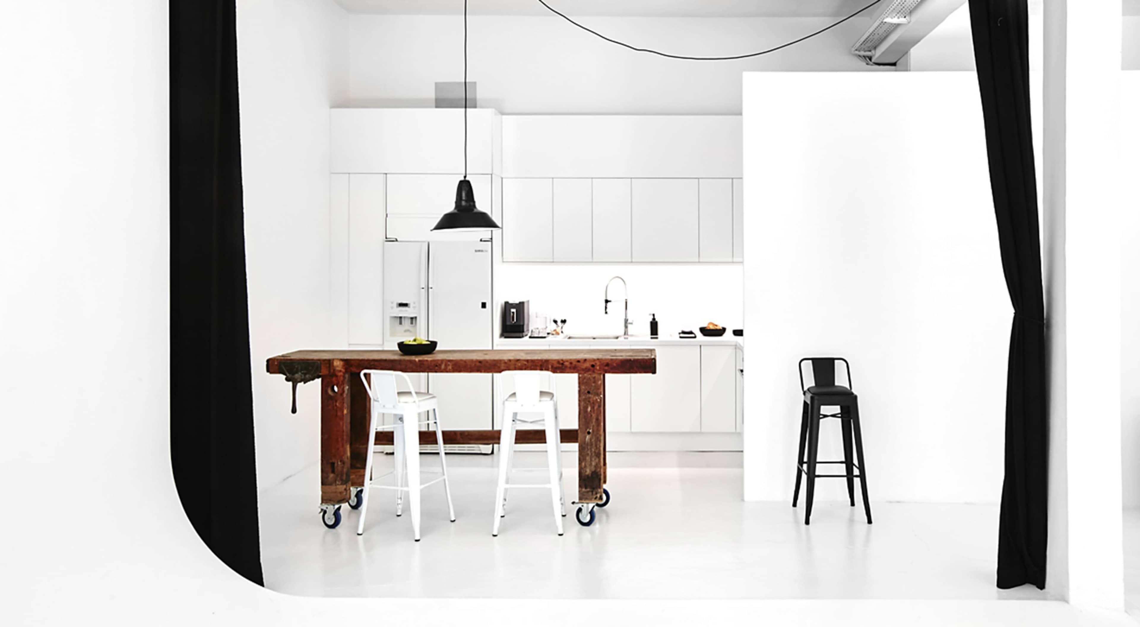 A minimalist kitchen with a wooden island and white cabinetry, set against a backdrop of white walls and black curtains.