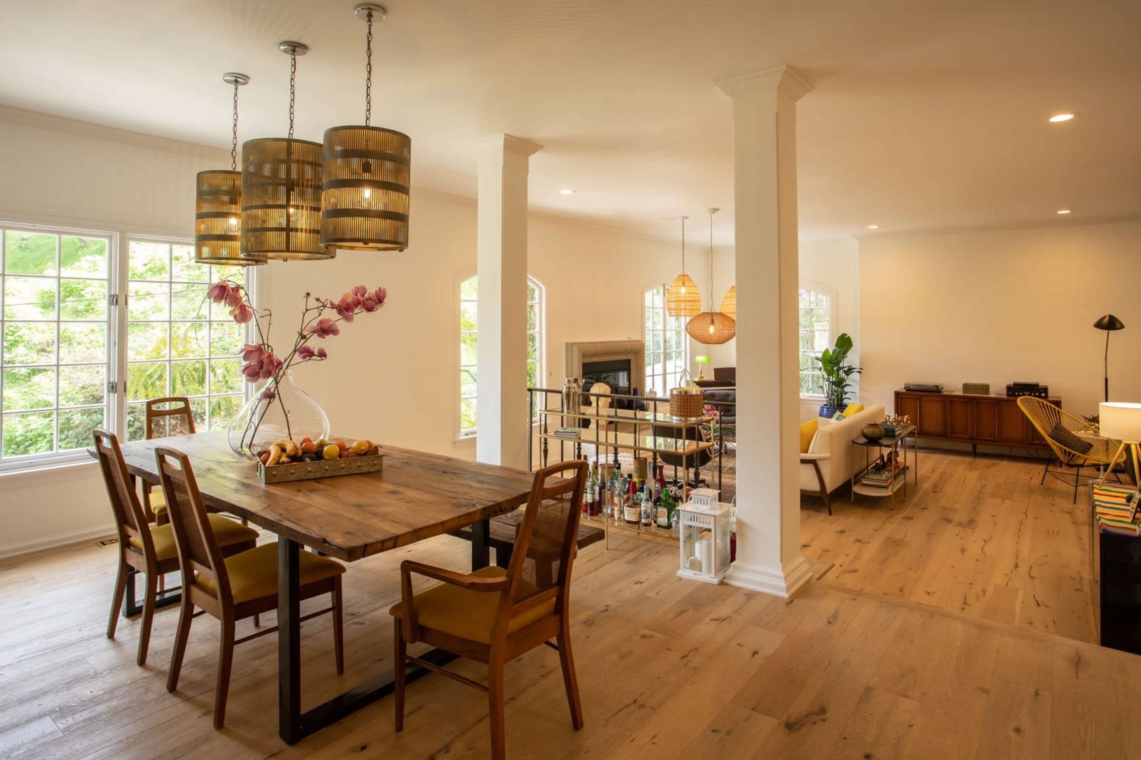 A dining area features a wooden table surrounded by chairs, with pendant lights above and a view of a sitting area with a fireplace in the background.