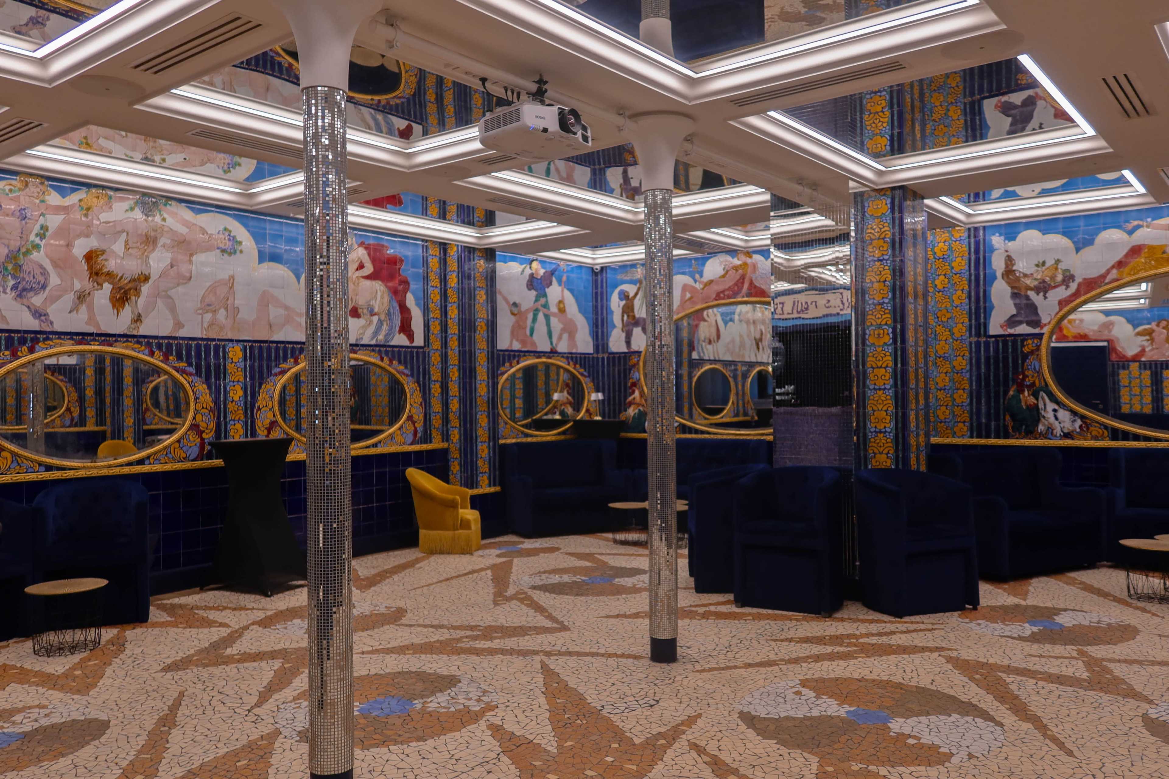 The interior features a spacious room with mosaic flooring, ornate murals on the walls, and a mix of blue velvet seating and mirrored accents.