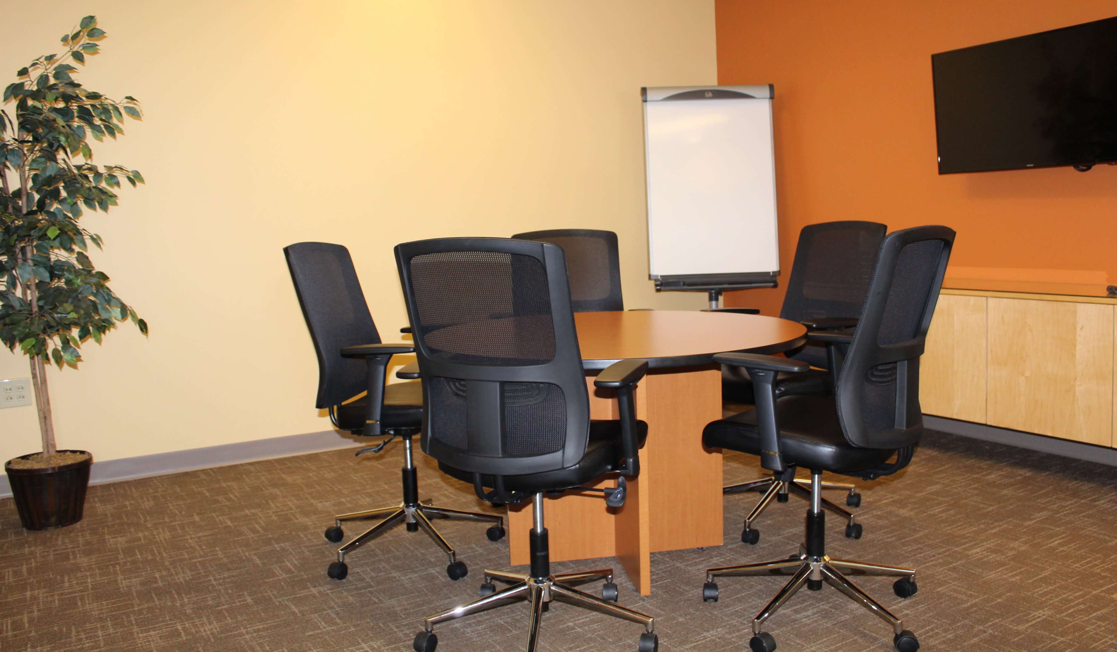 A conference room features a round table surrounded by five black office chairs, with a flip chart, a TV, and a potted plant in the background.