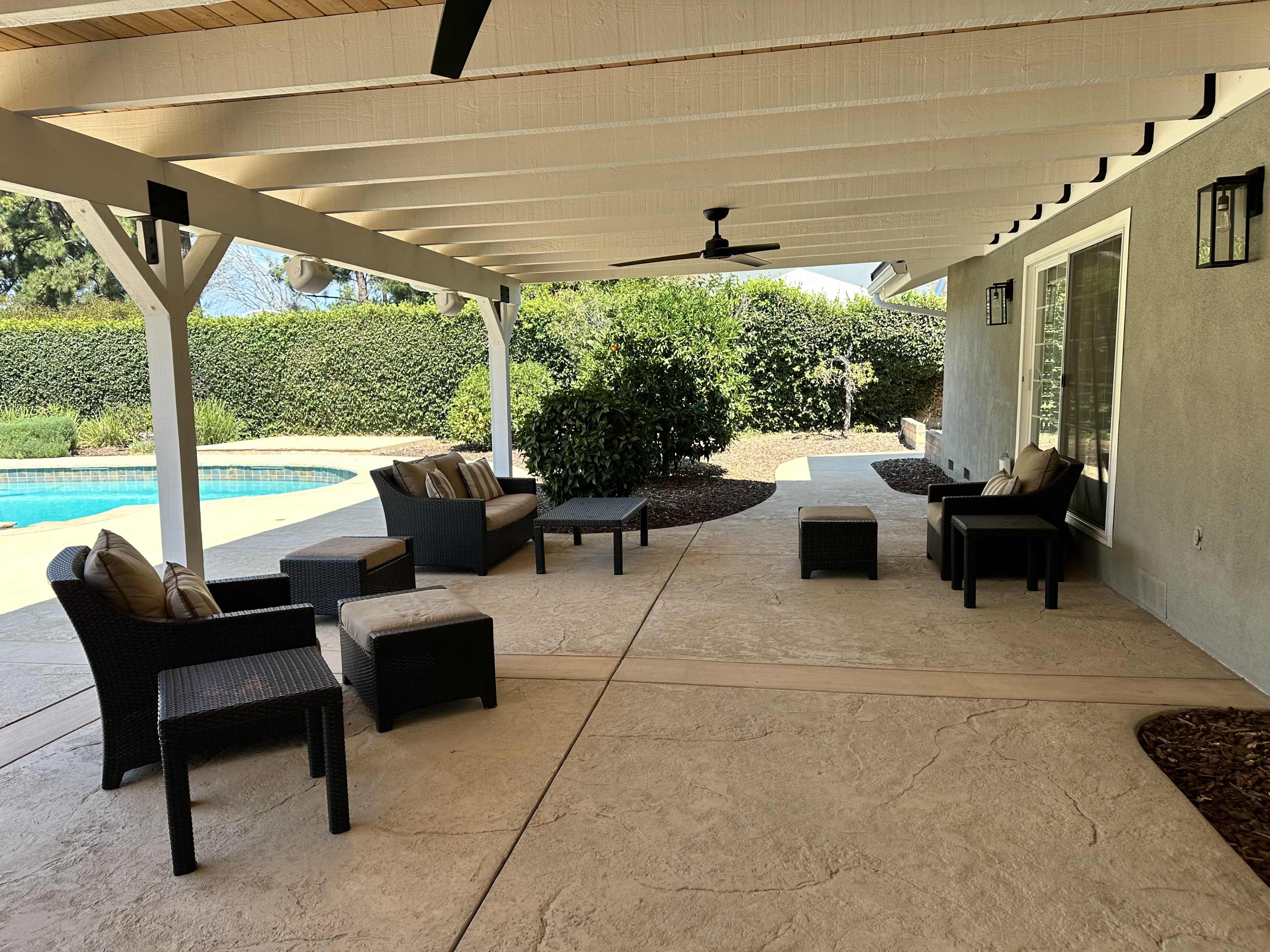 A shaded outdoor patio features several wicker seating options surrounding a small table near a swimming pool.