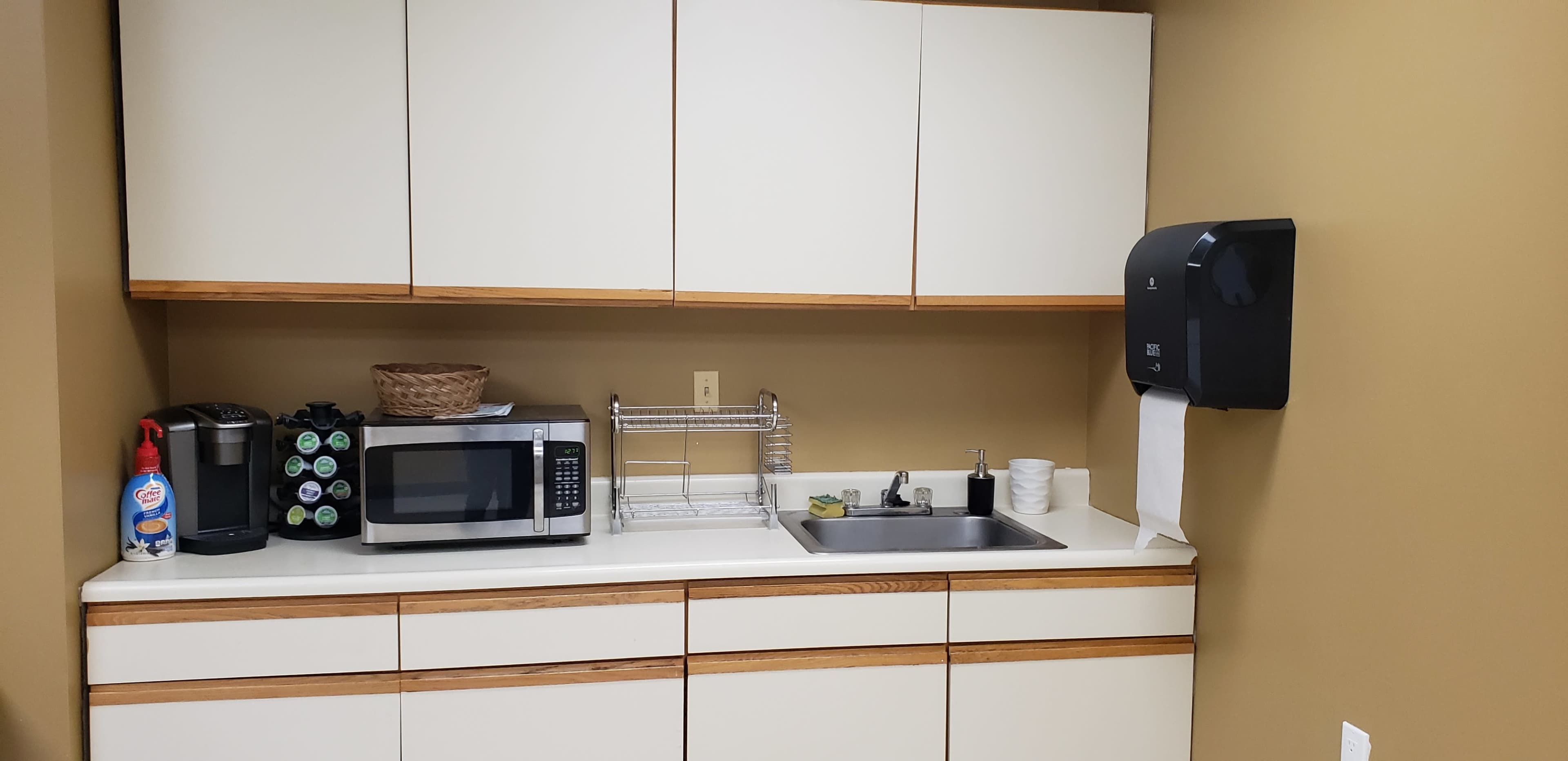 A break room kitchen area with a microwave, a coffee maker, a sink, and a paper towel dispenser against a brown wall.