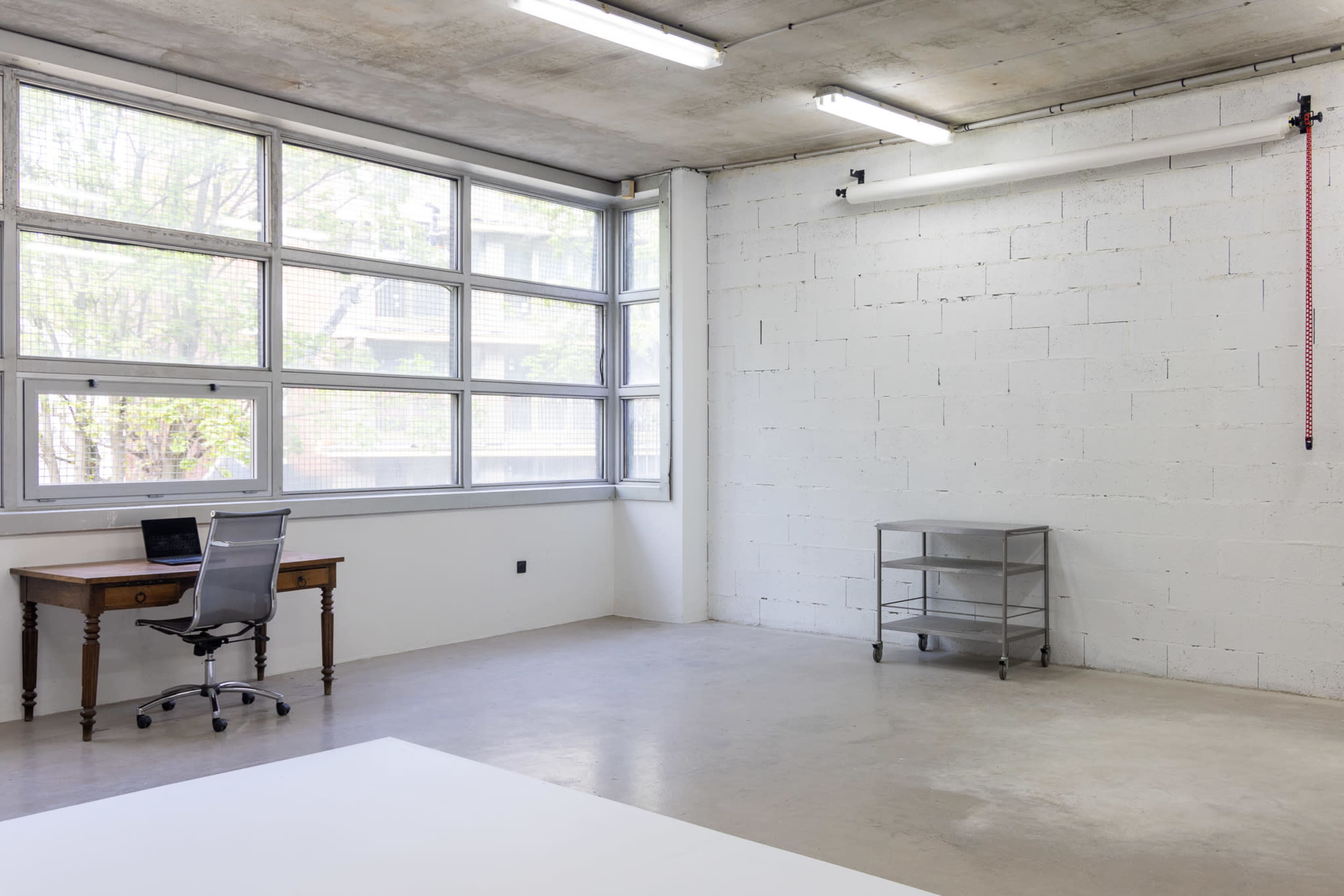 The image shows a spacious, minimally furnished office with a desk and chair near a large window, and a metal shelving unit against a bare wall.
