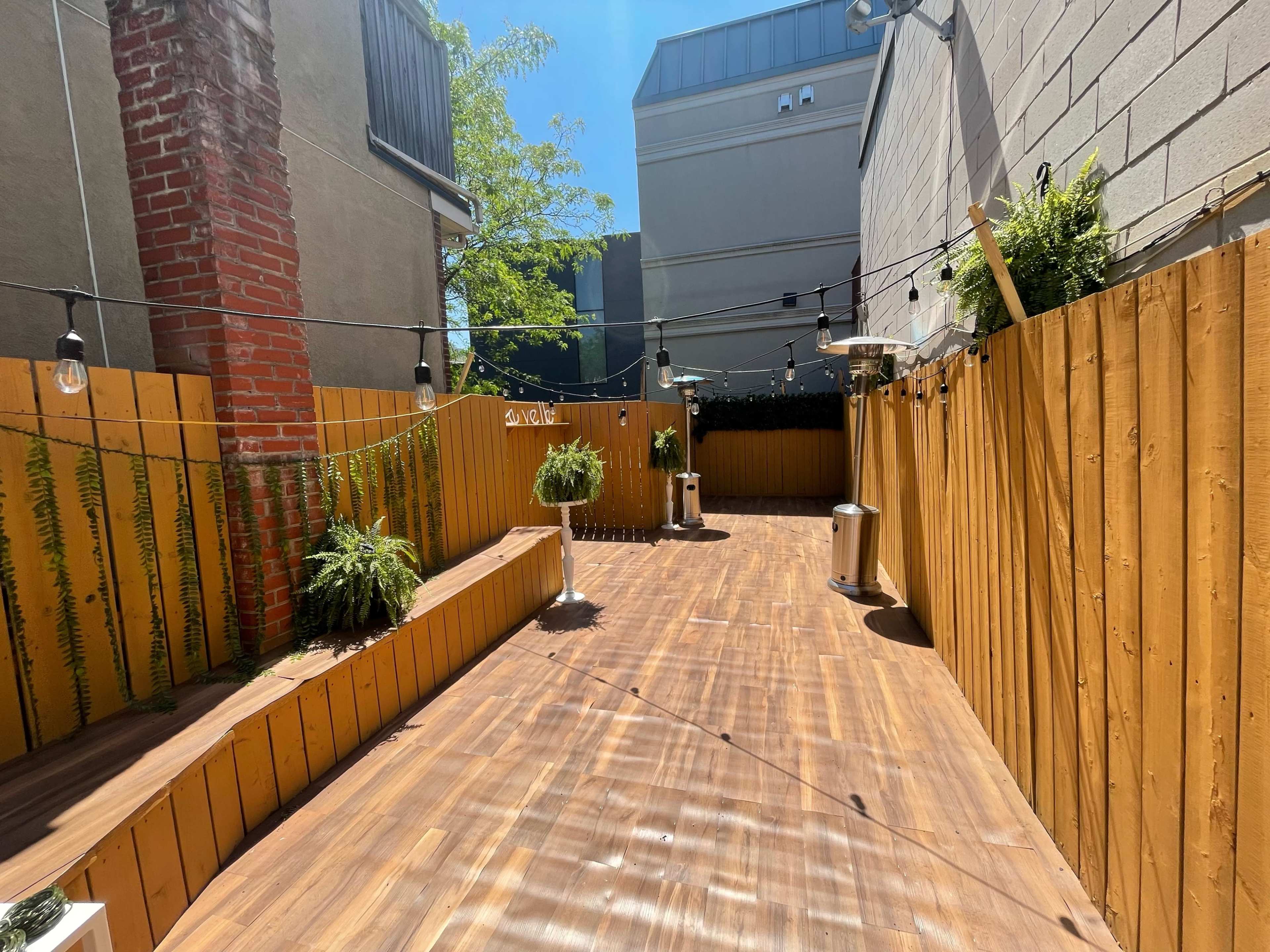 A narrow, sunlit outdoor patio features wooden decking, enclosed by wooden fencing and adorned with potted plants and hanging lights.