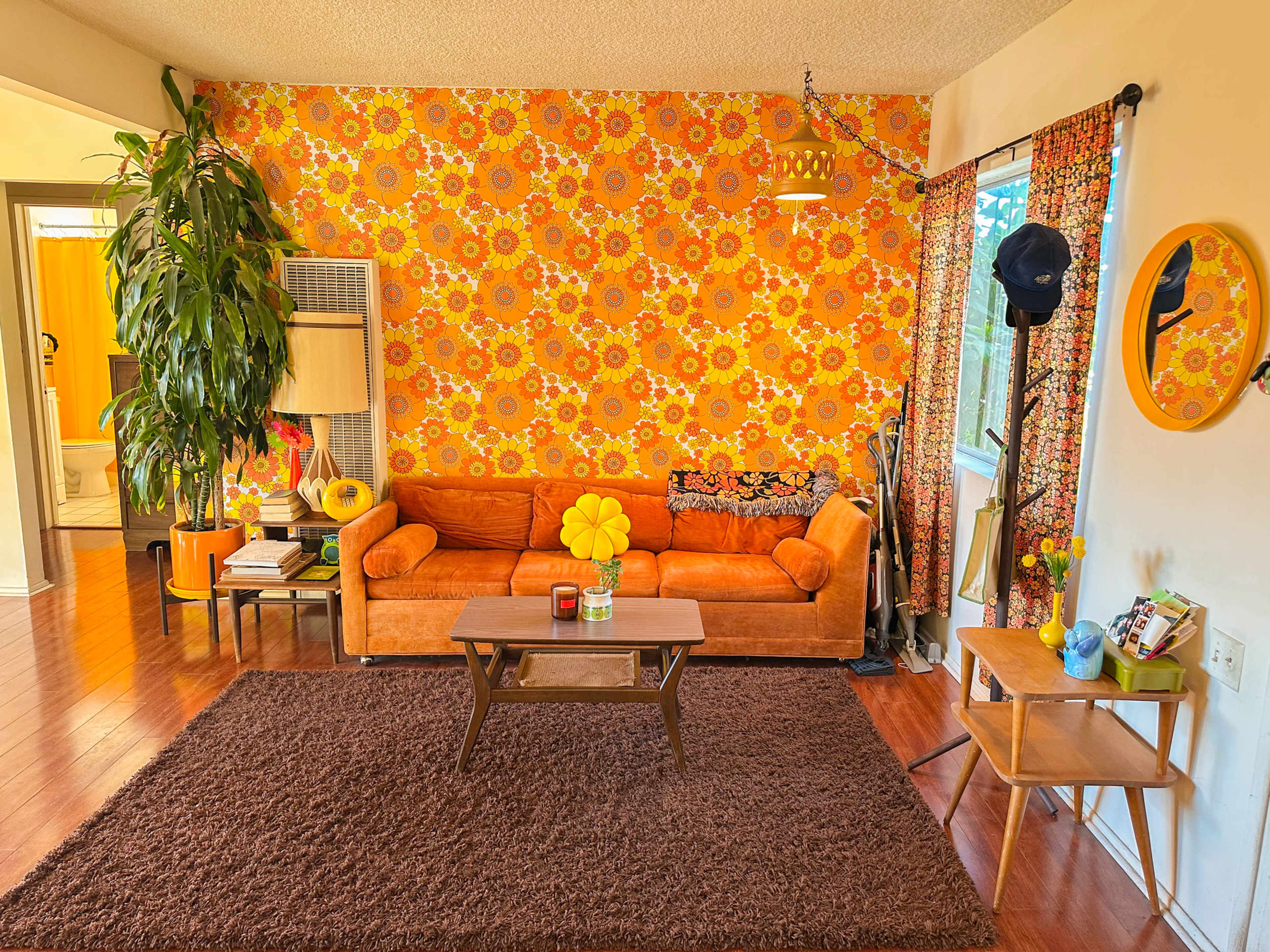 The image shows a vintage living room featuring a bright orange couch, a brown rug, a patterned yellow wallpaper with flowers, and various decorative items on a coffee table and side table.