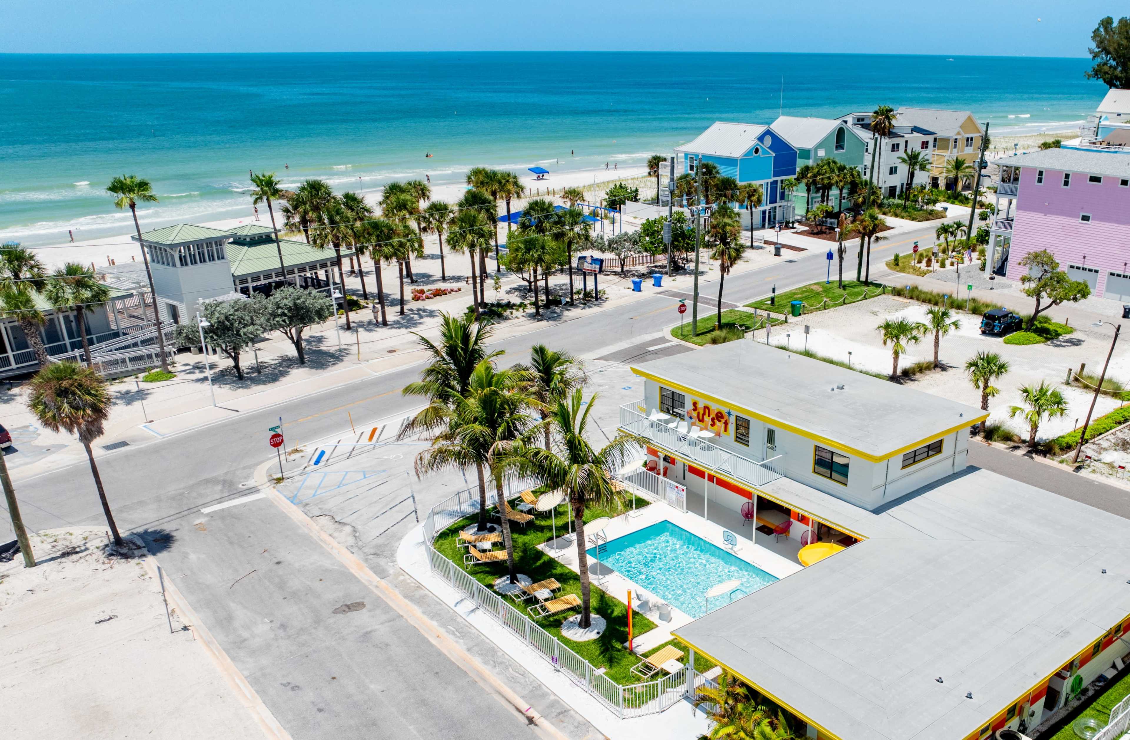 The image shows a beachfront scene featuring a hotel with a swimming pool surrounded by palm trees, adjacent to a sandy beach and colorful beach houses.