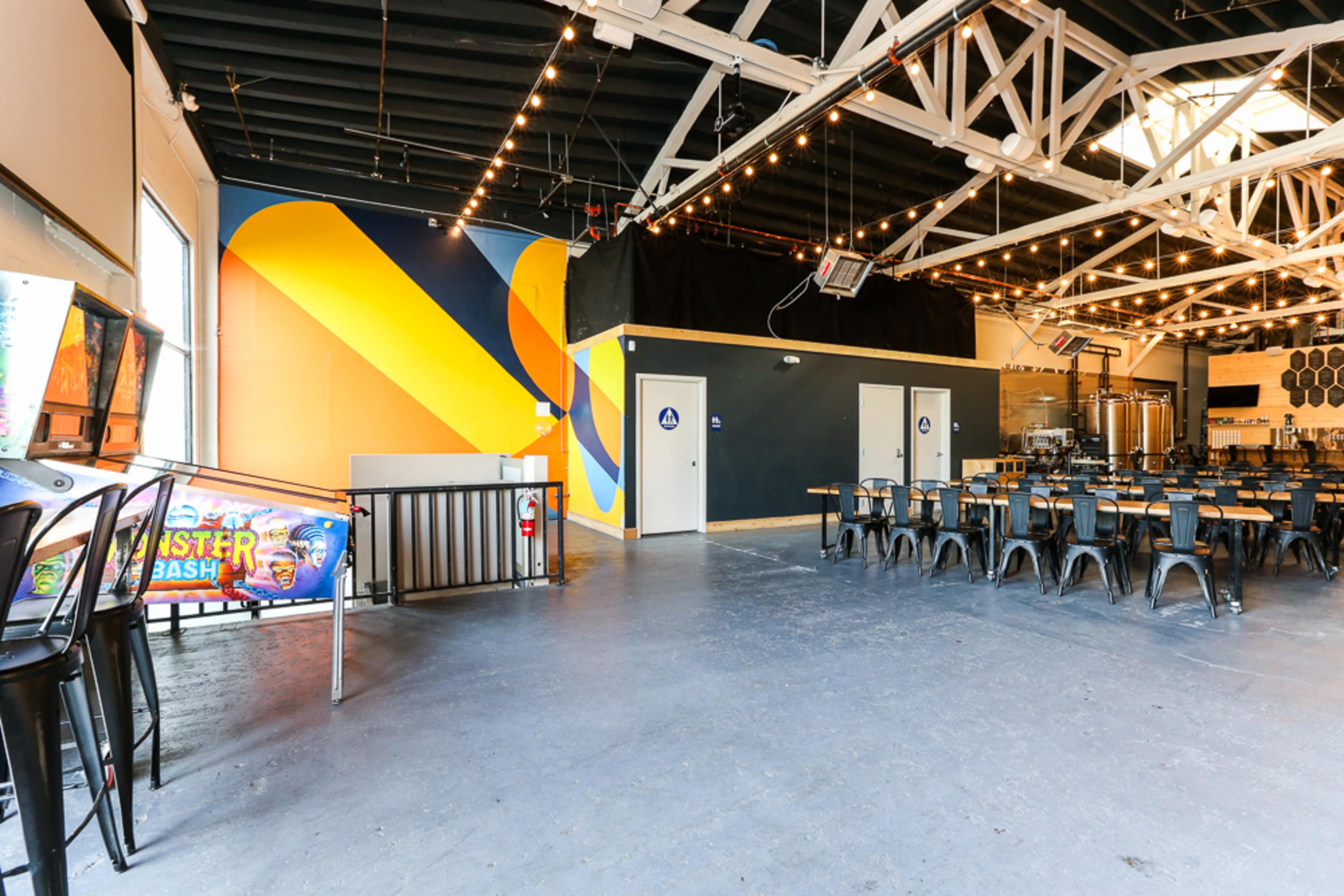 A spacious interior of a venue featuring colorful wall art, seating arrangements of metal chairs and tables, and a bar area.