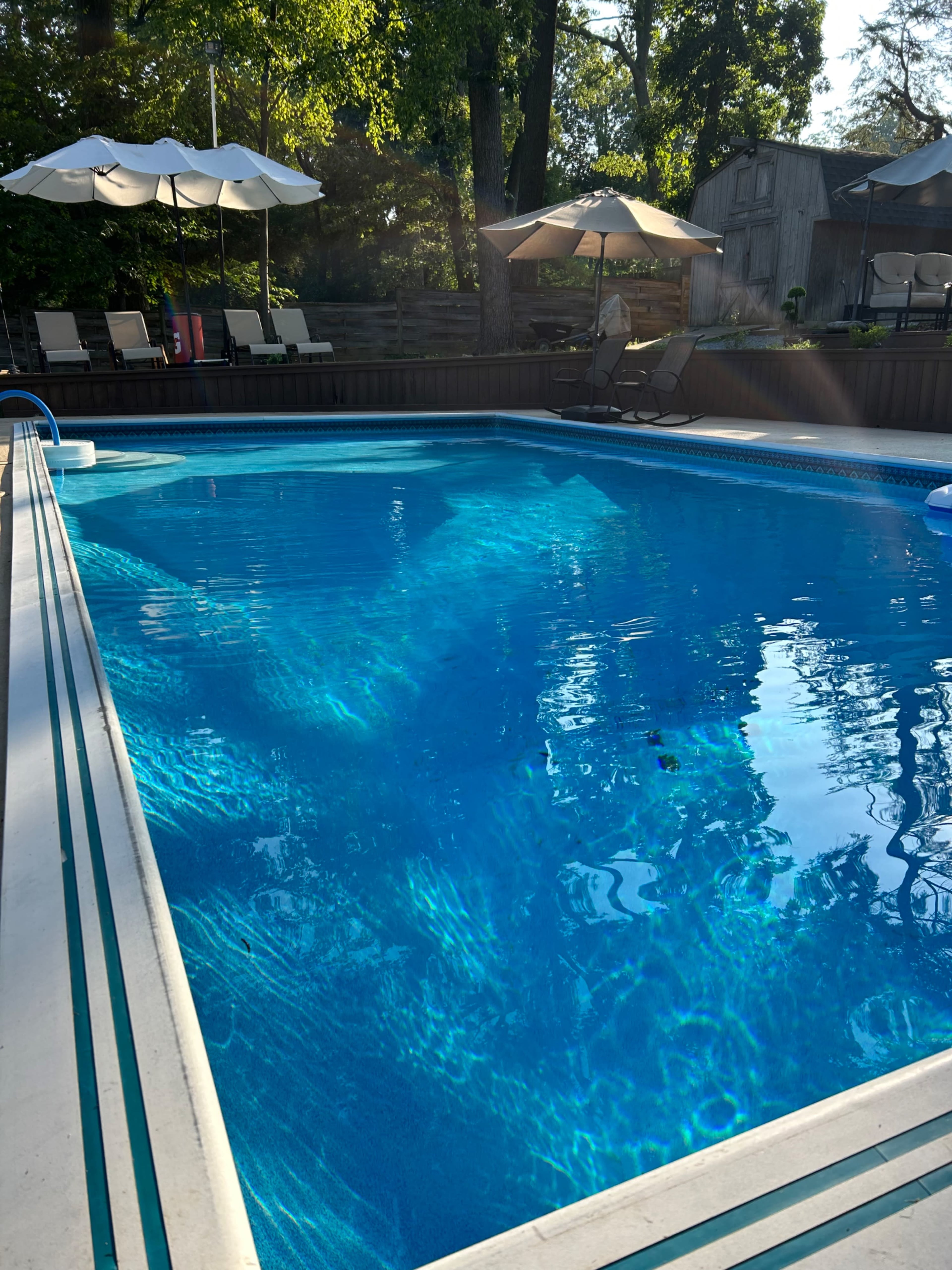 A clear blue swimming pool is surrounded by lounge chairs and umbrellas in a shaded outdoor area.