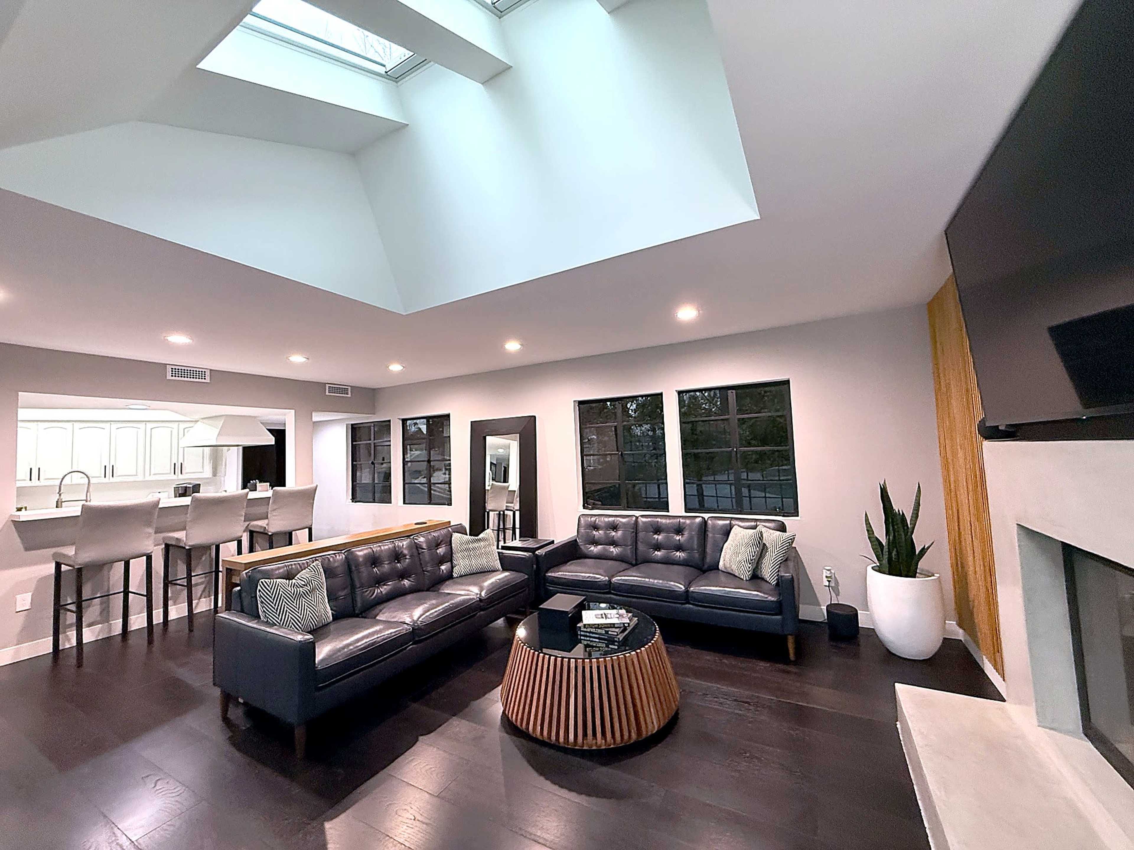 The image features a modern living room with two black leather sofas, a round wooden coffee table, and large windows allowing natural light to enter.