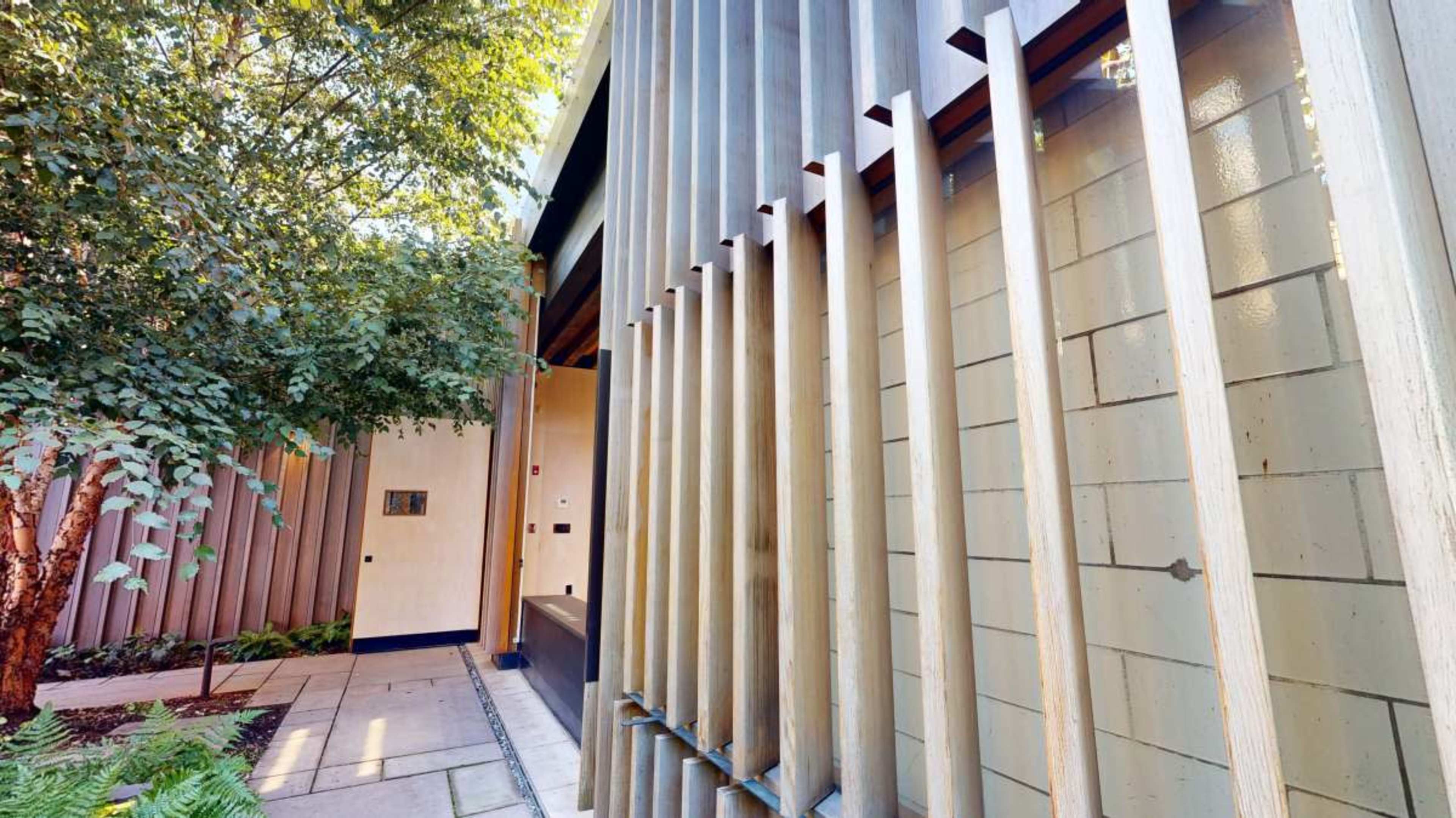 The image shows a modern architectural entrance featuring vertical wooden slats and a landscaped pathway with plants.
