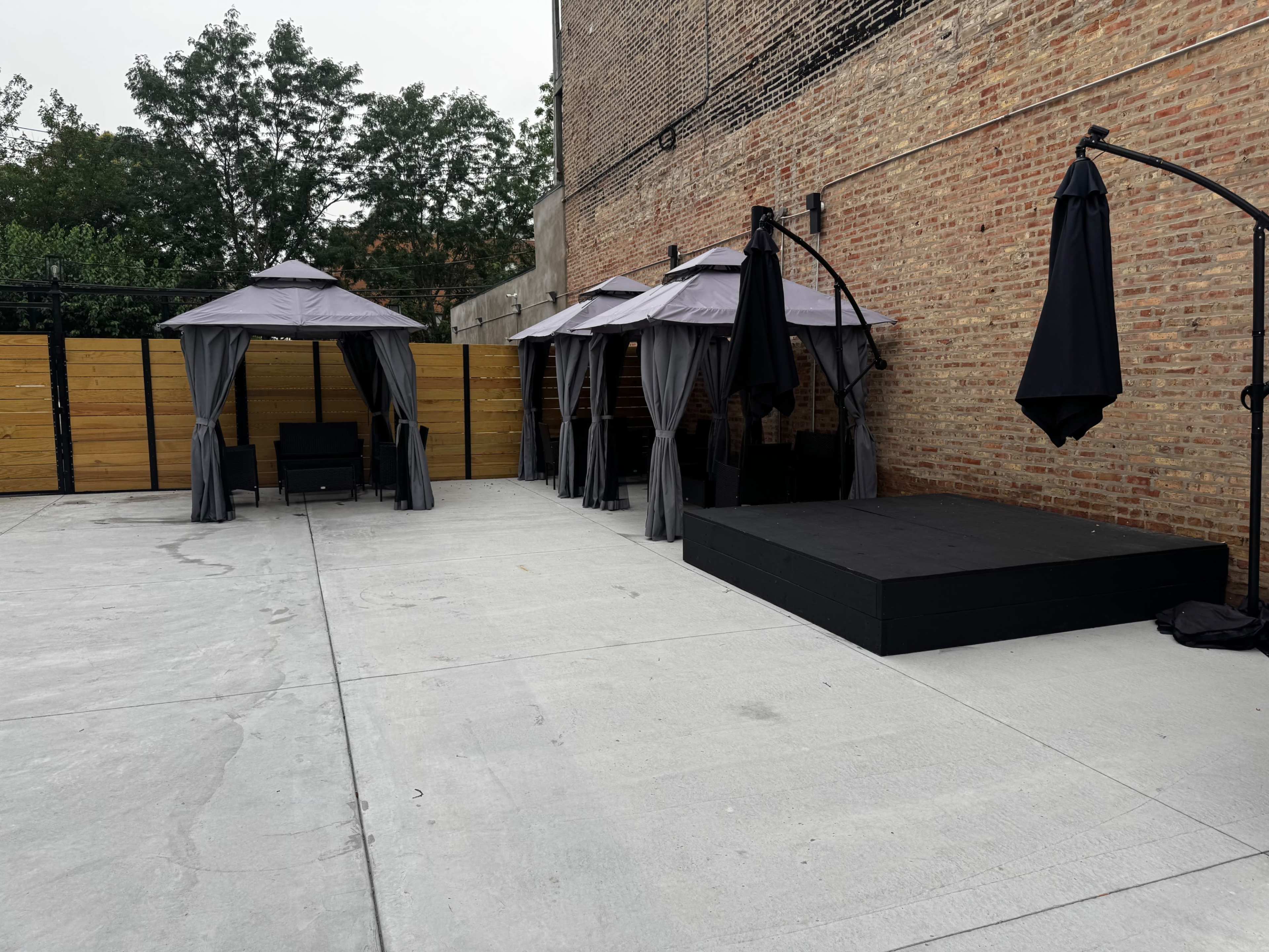 The image shows a patio area featuring several canopies, a raised platform, and wooden fencing against a brick wall.