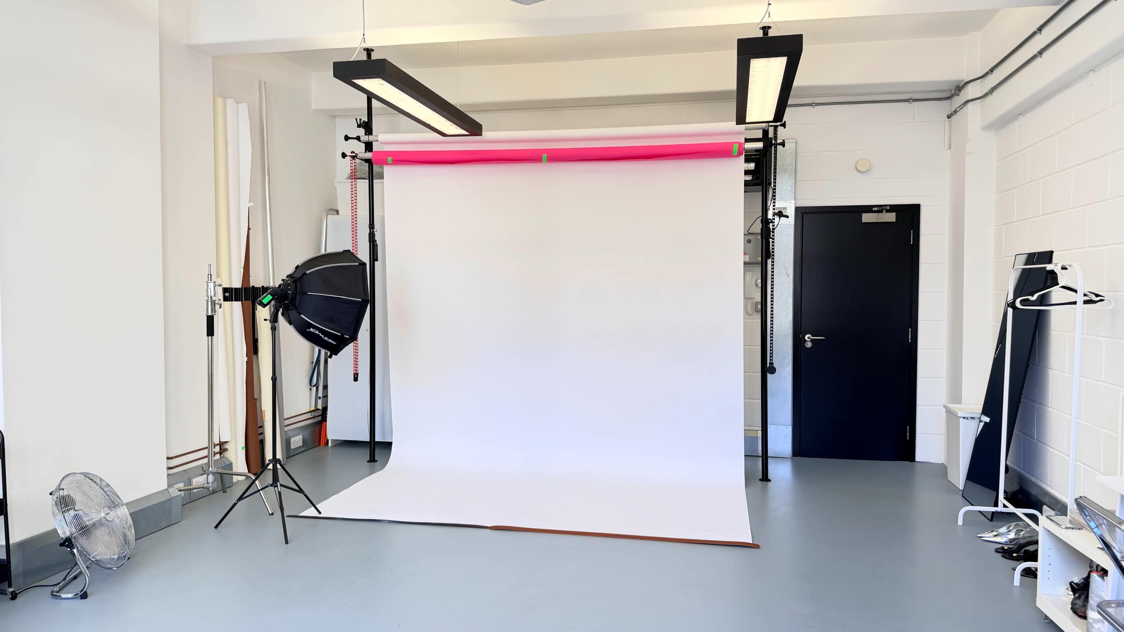 The image shows a photography studio with a white backdrop, lighting equipment, and a few accessories arranged against plain walls.