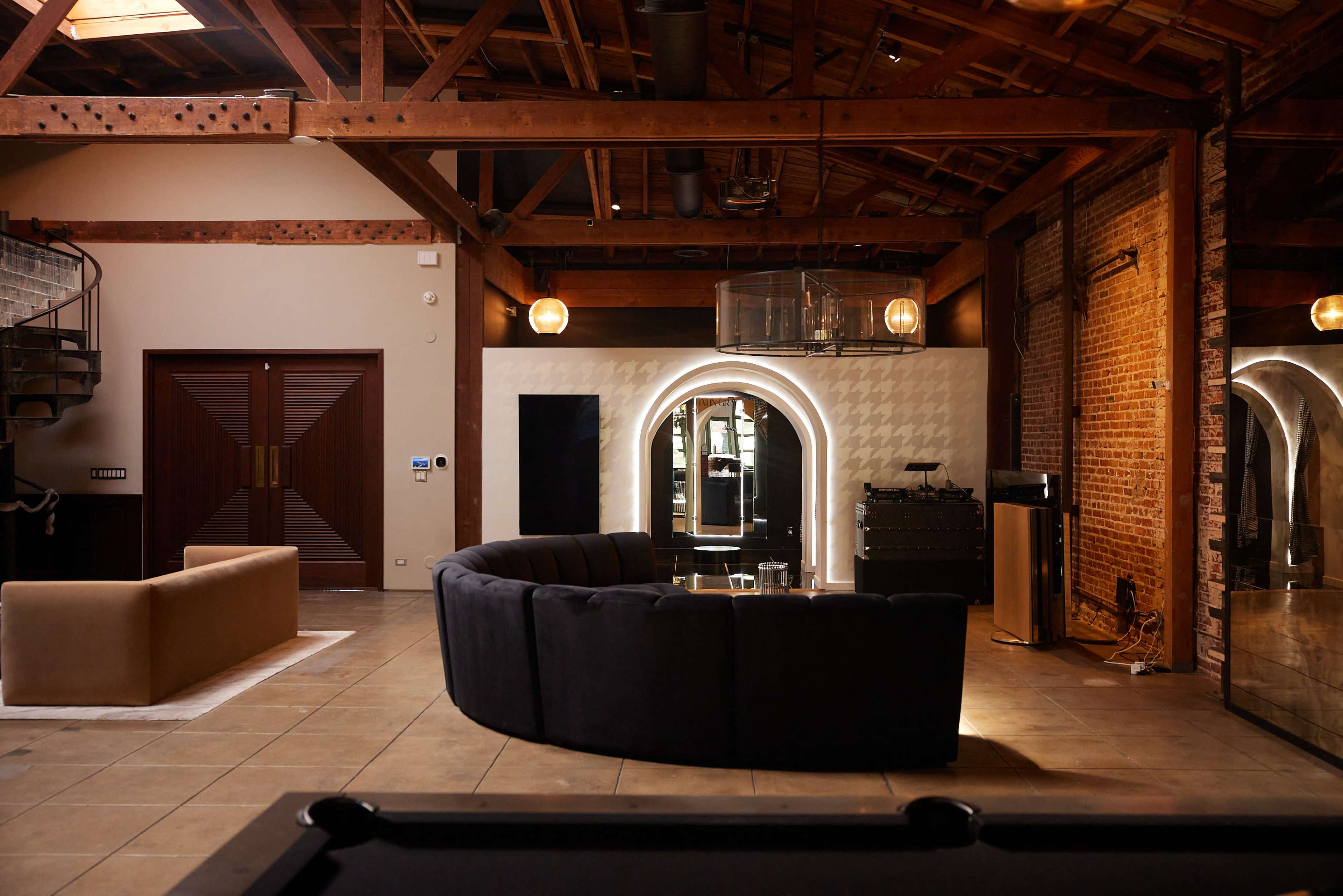 The image shows a spacious interior with a circular dark sofa, a beige couch, and exposed brick walls under a wooden ceiling with industrial lighting fixtures.