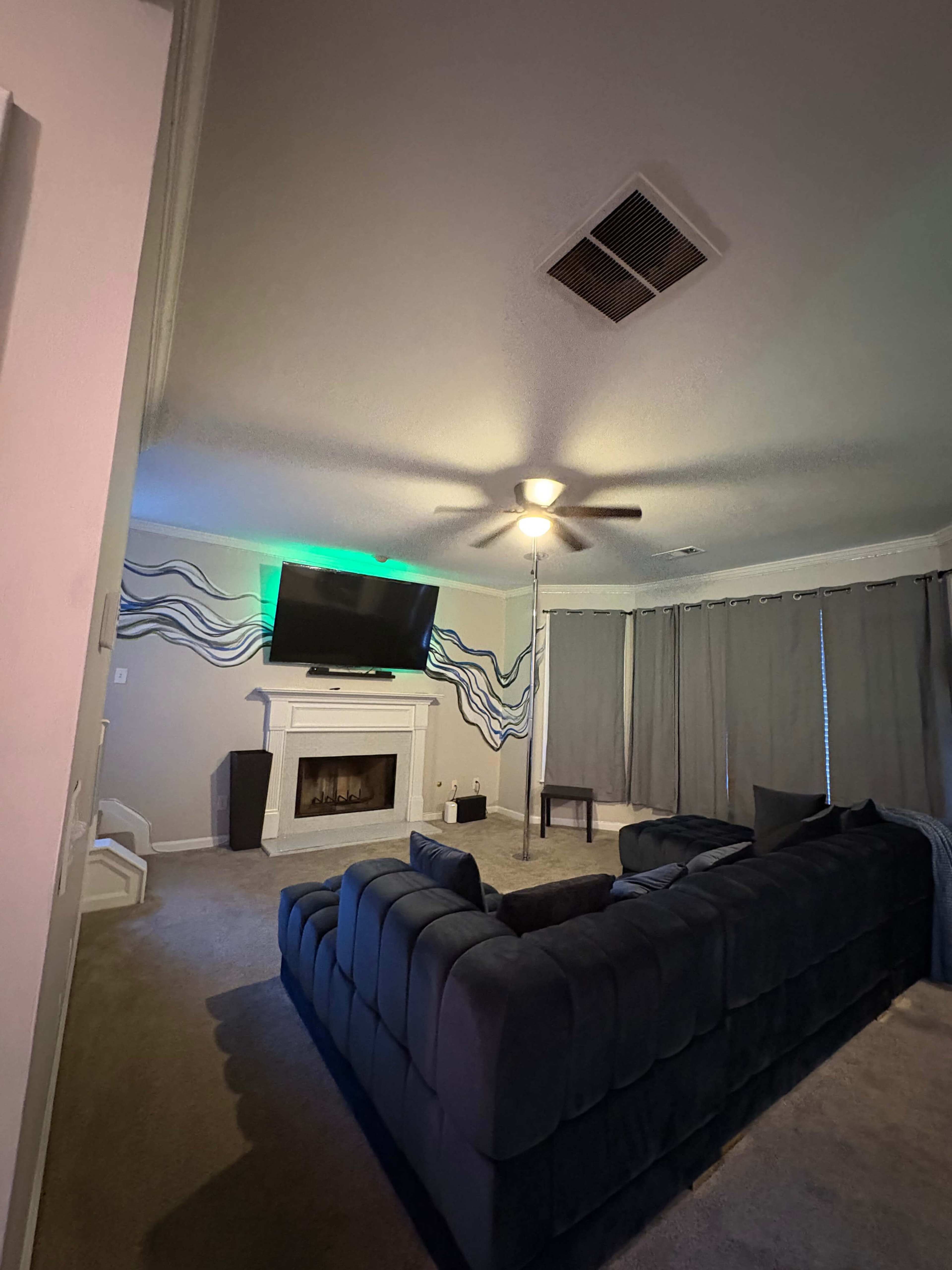 A living room features a sectional sofa, a mounted television above a fireplace, and curtains partially drawn at a window.