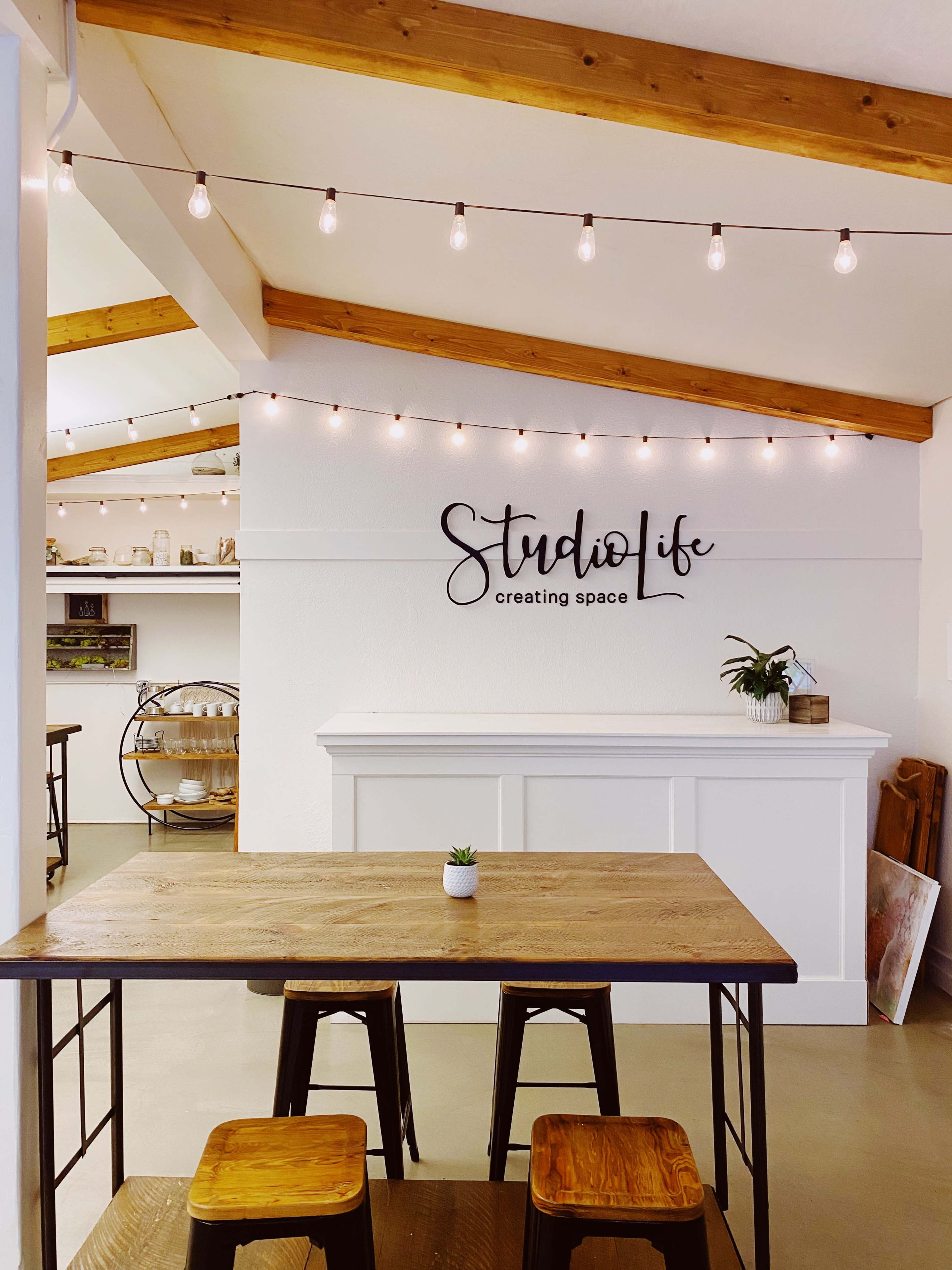 A bright interior space with a wooden table, metal stools, and a sign reading "StudioLife" on the wall, decorated with string lights above.