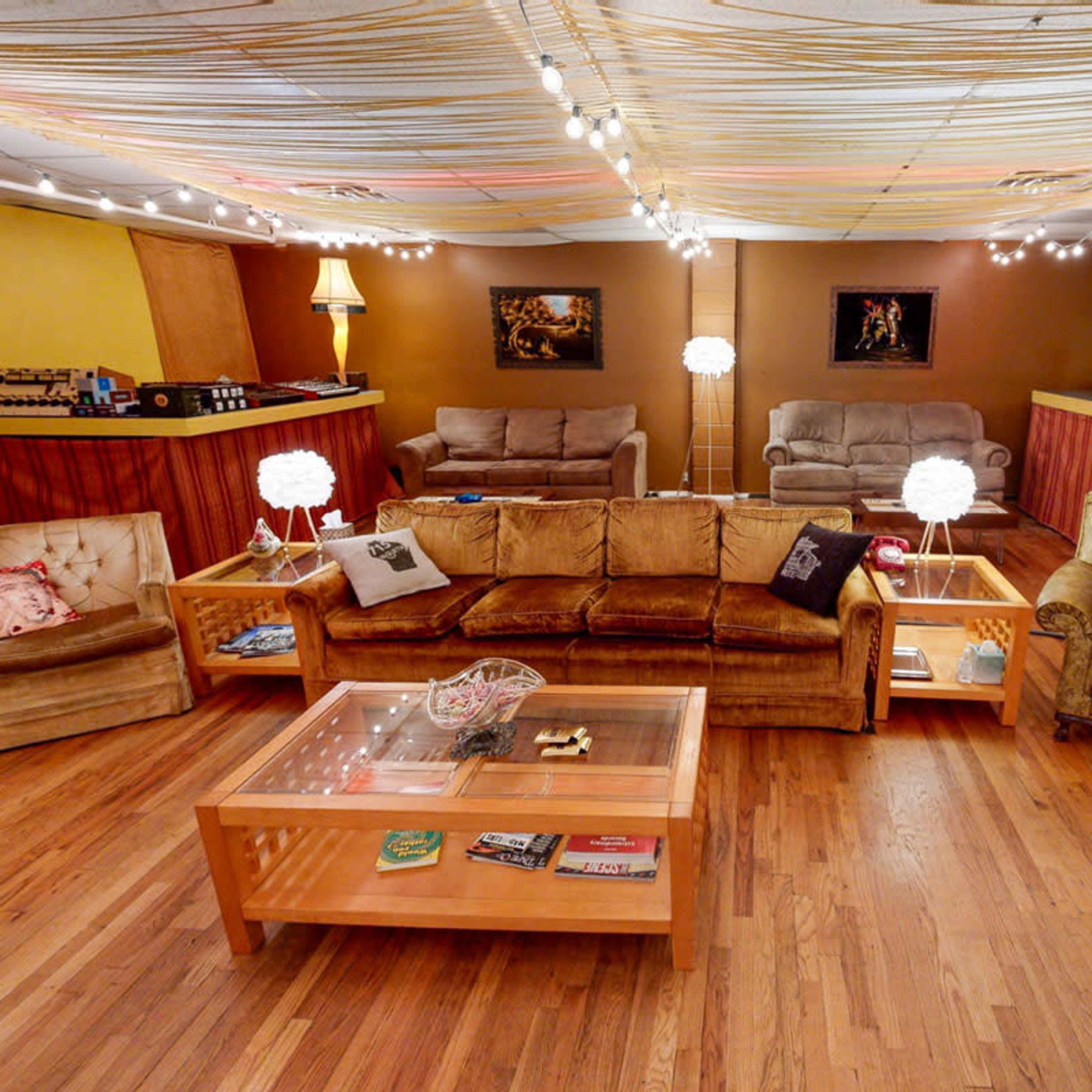 The image shows a cozy lounge area with multiple seating arrangements, wooden flooring, a glass coffee table, and soft lighting from hanging lamps and string lights.