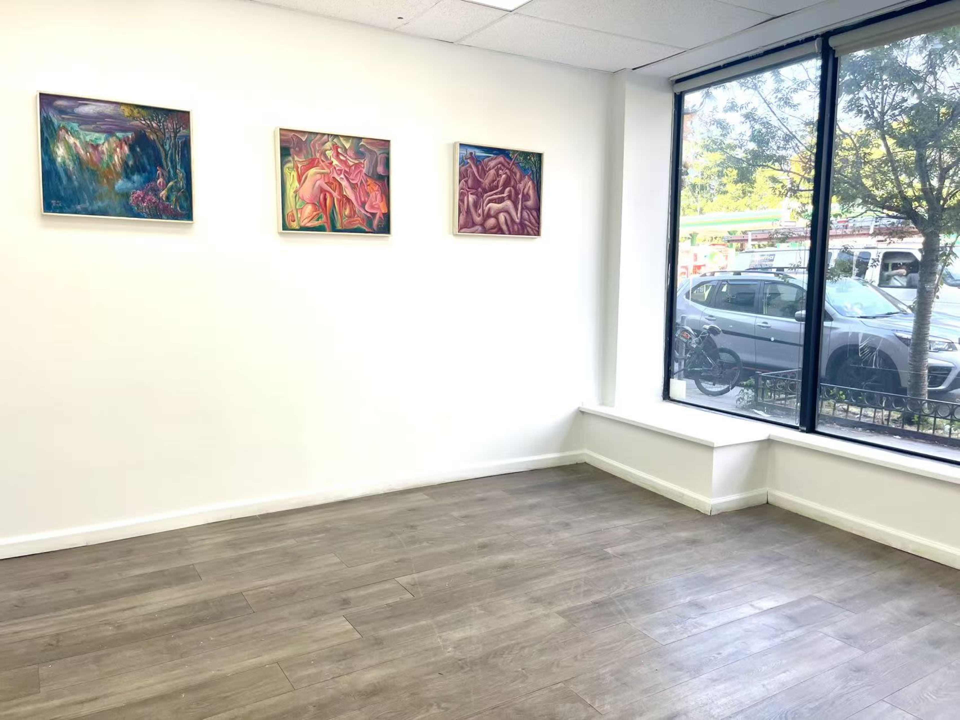 The image shows an empty art gallery with three paintings hanging on a white wall beside a large window that overlooks a street with parked cars.