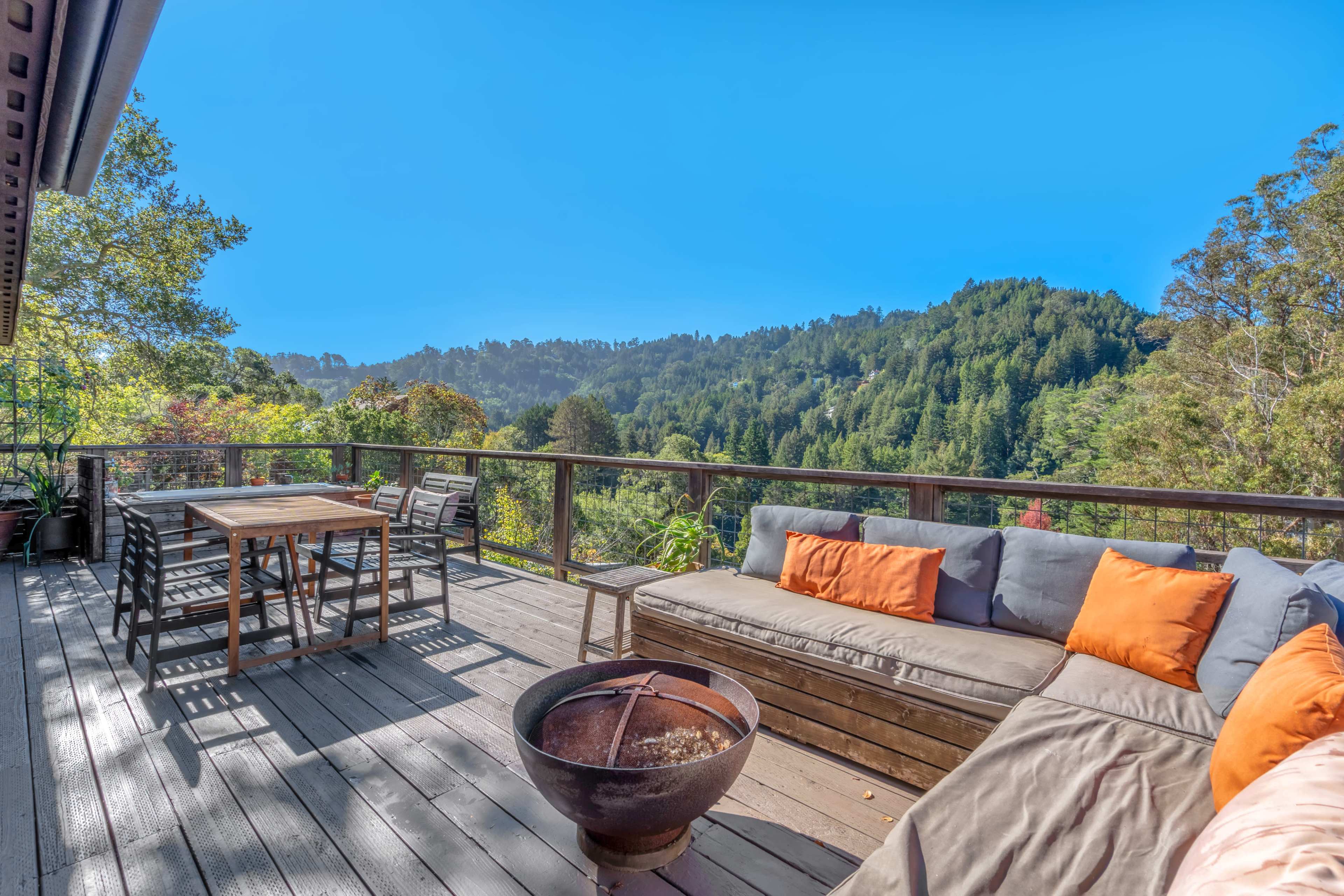 A spacious wooden deck features a fire pit, a dining table, and a sectional sofa with orange cushions, overlooking a scenic landscape of trees and hills.