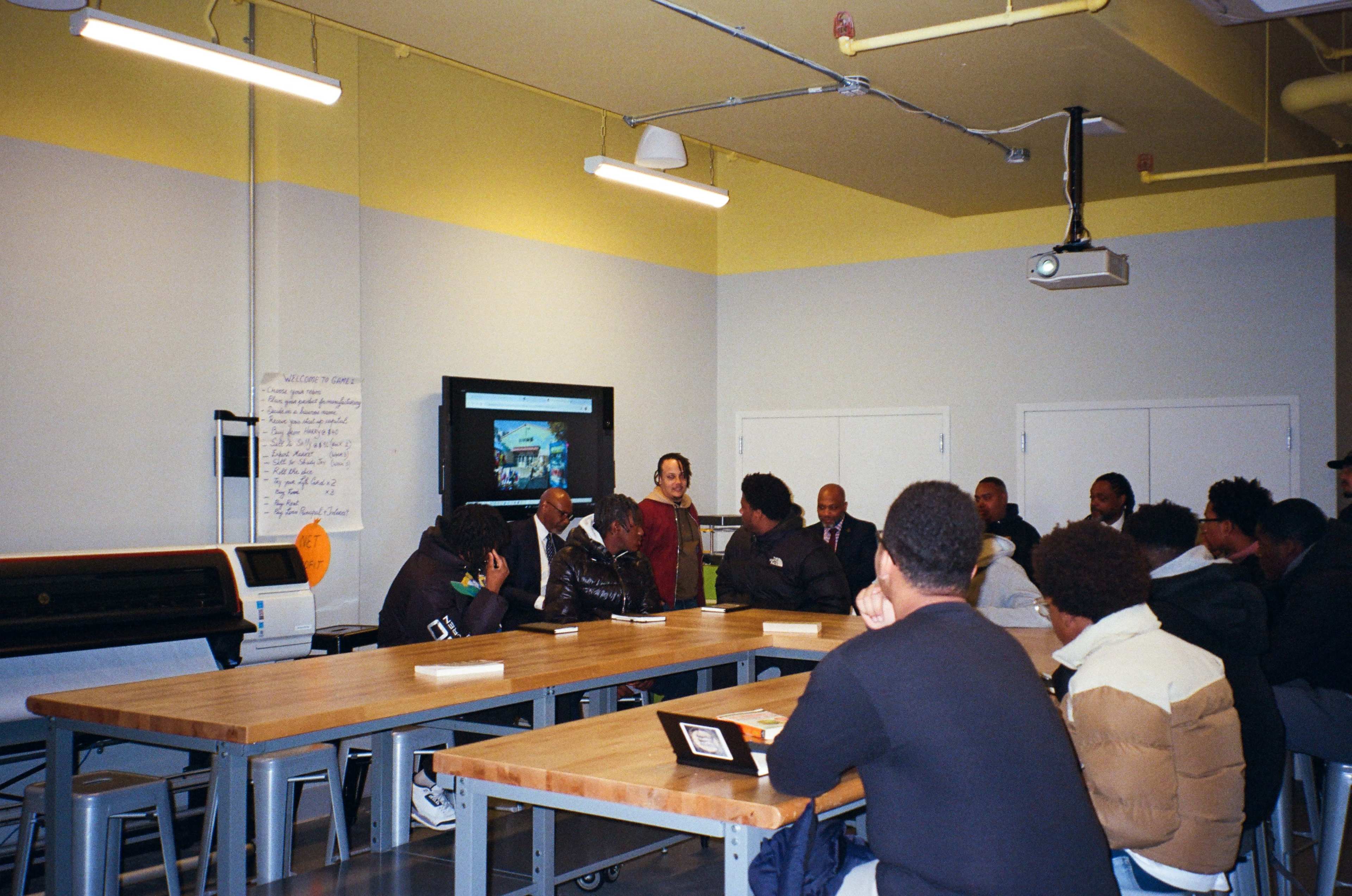 A group of people is sitting around a large table in a classroom setting, engaged in a discussion while a person stands and speaks to them.