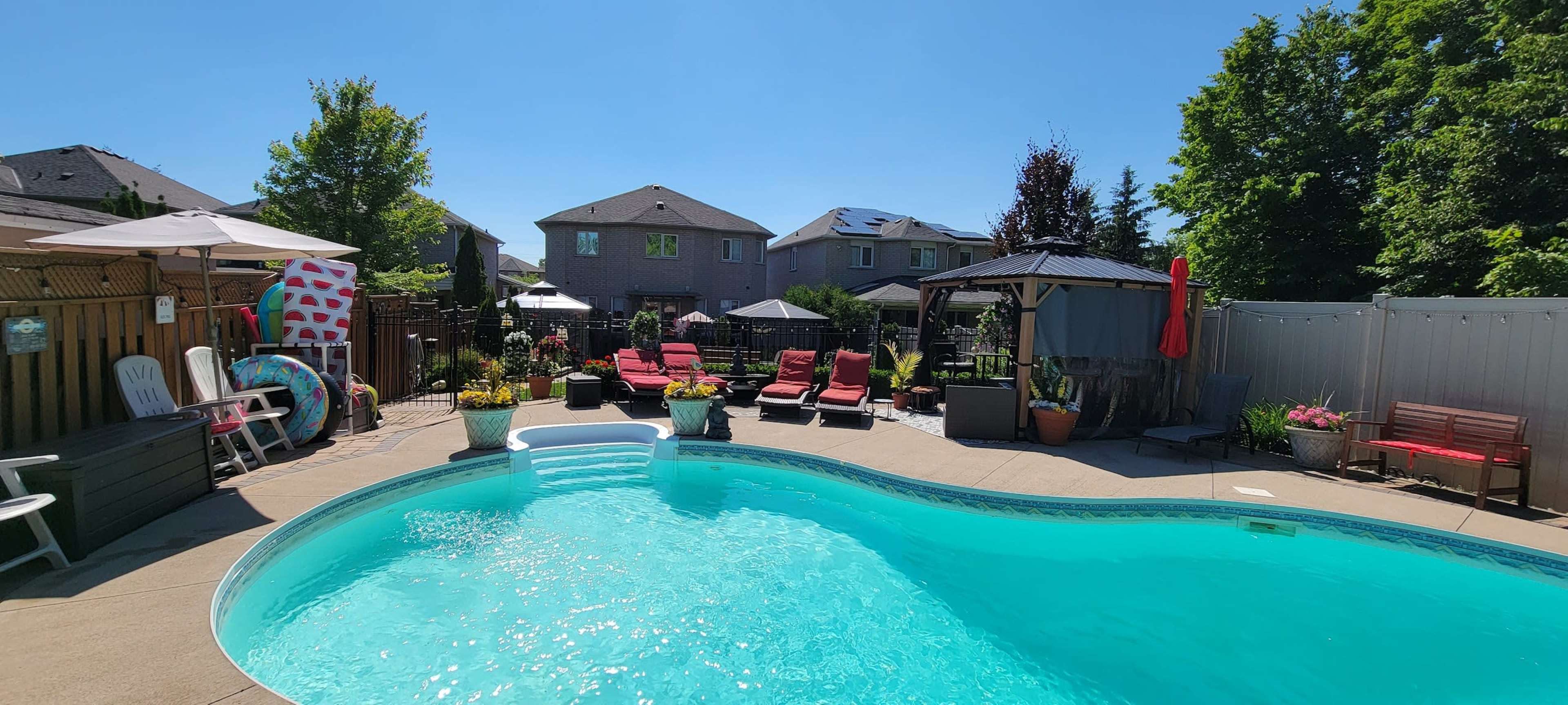 A backyard swimming pool surrounded by lounge chairs, potted plants, and a gazebo, with residential houses in the background.