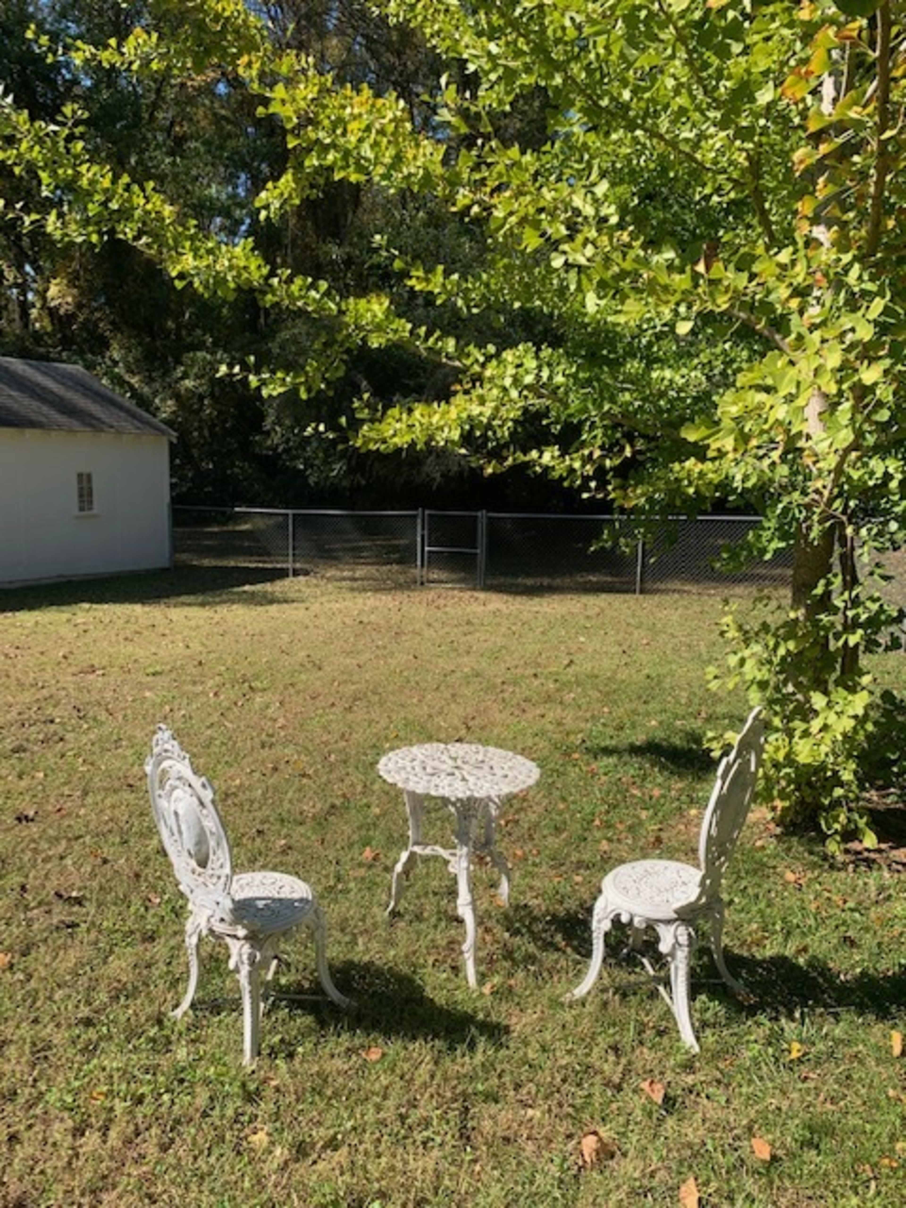 A small white metal table with three matching chairs is placed on a grassy area surrounded by trees and a fence.