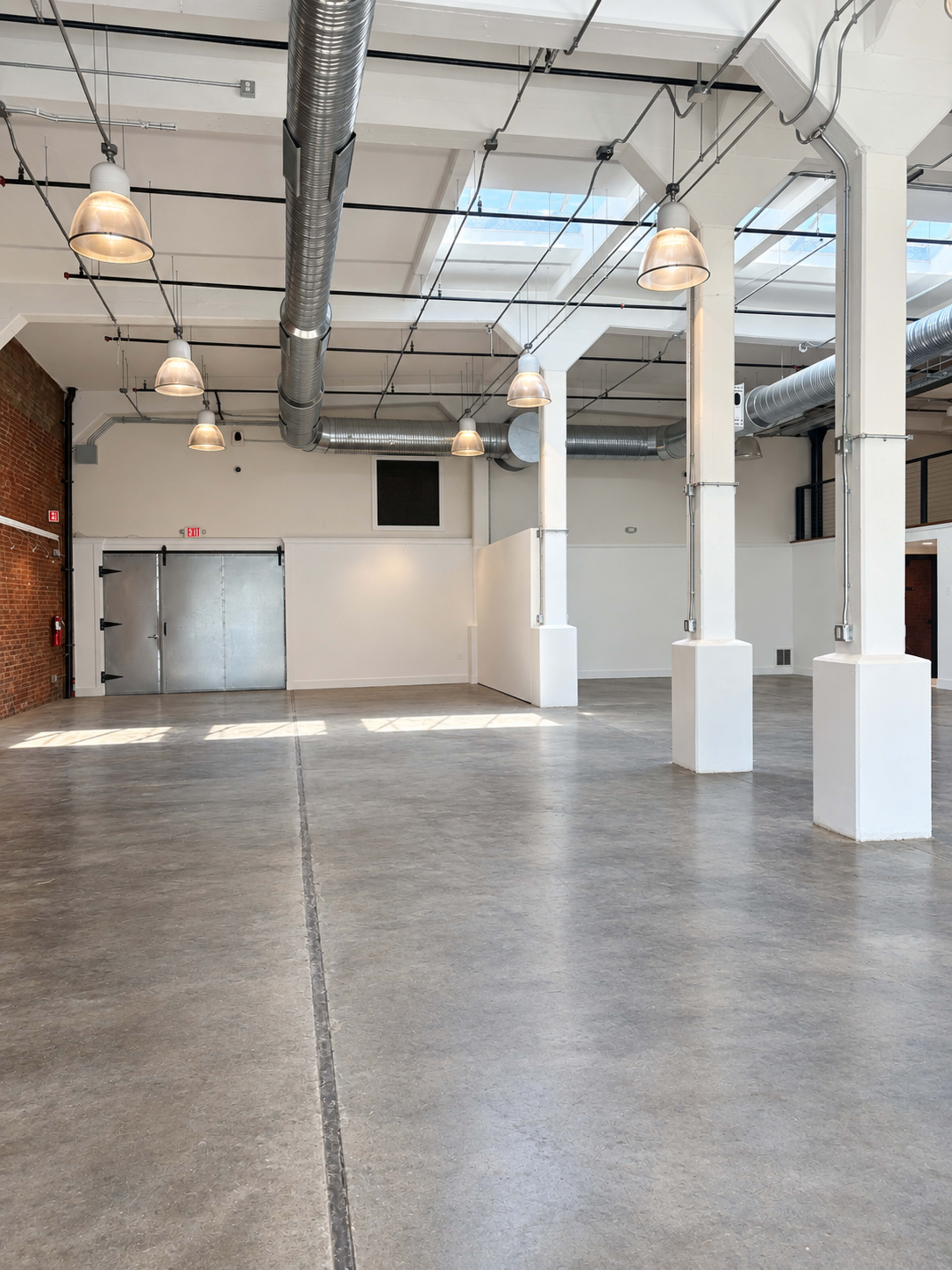 The image shows a large, empty industrial space with concrete flooring, exposed ductwork, and several pendant lights hanging from the ceiling.