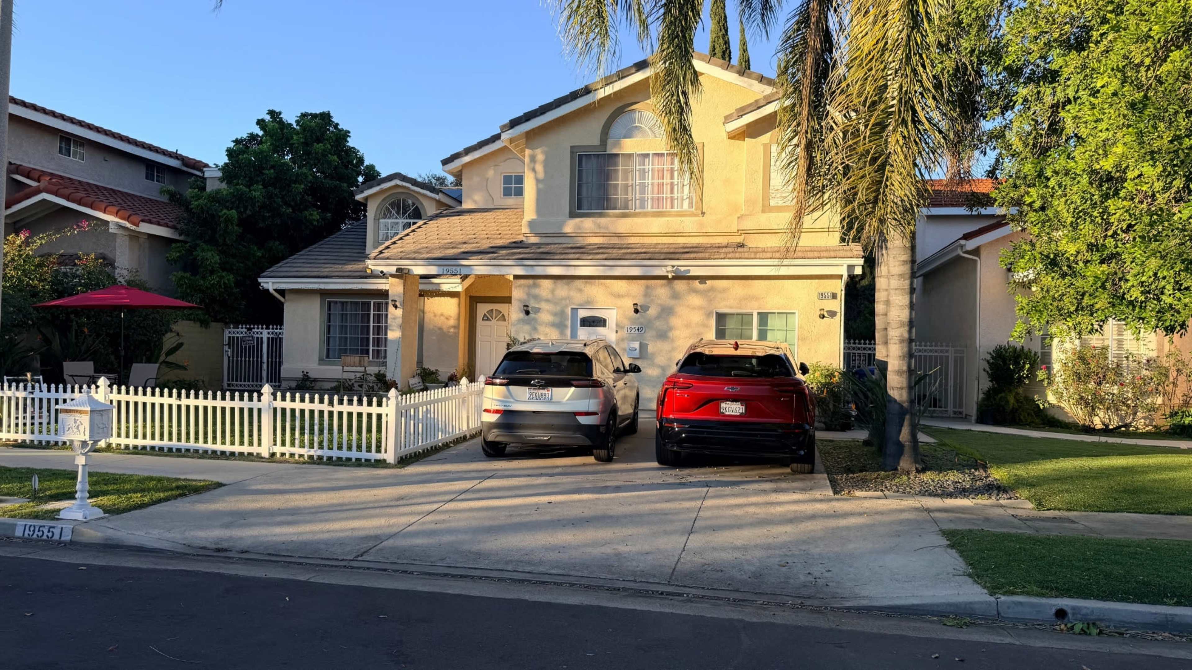 A two-story house with a white picket fence has two parked cars in the driveway, surrounded by palm trees and a clear blue sky.