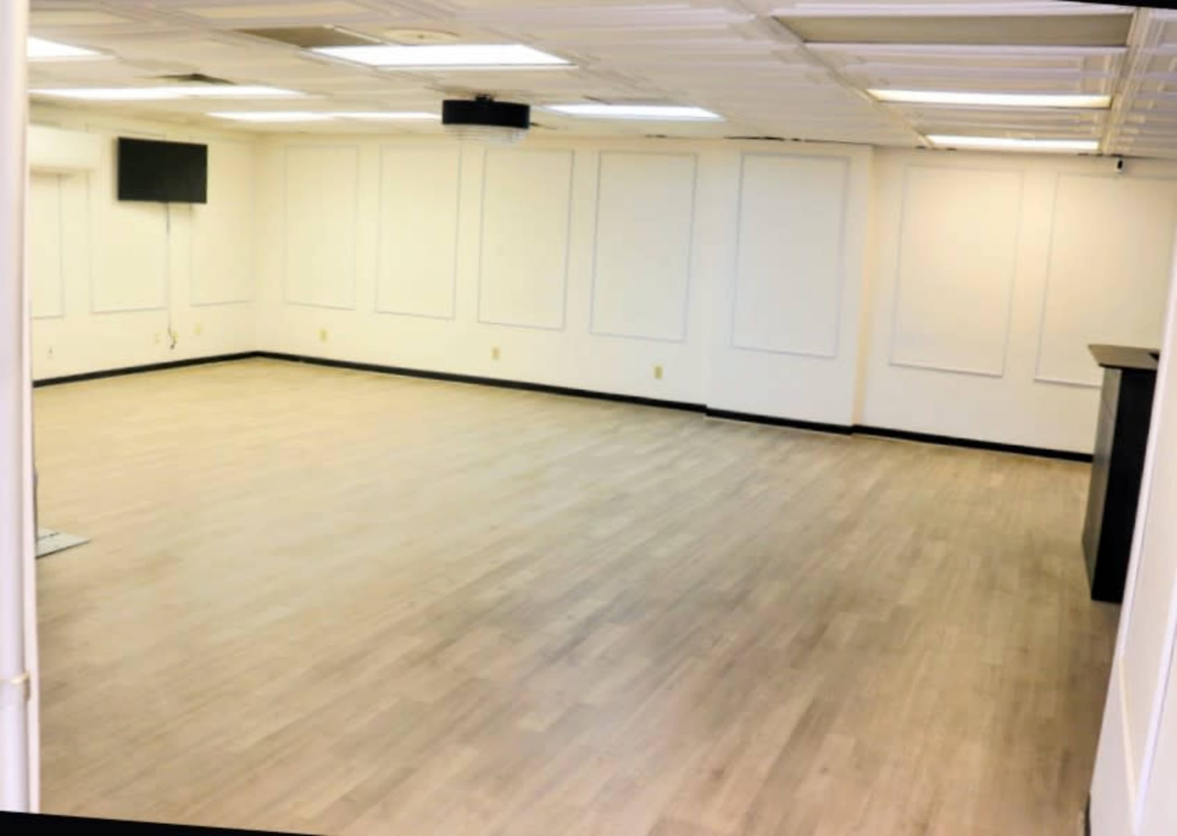 The image shows an empty, well-lit room with light-colored wooden flooring and white walls, featuring a small wall-mounted television.