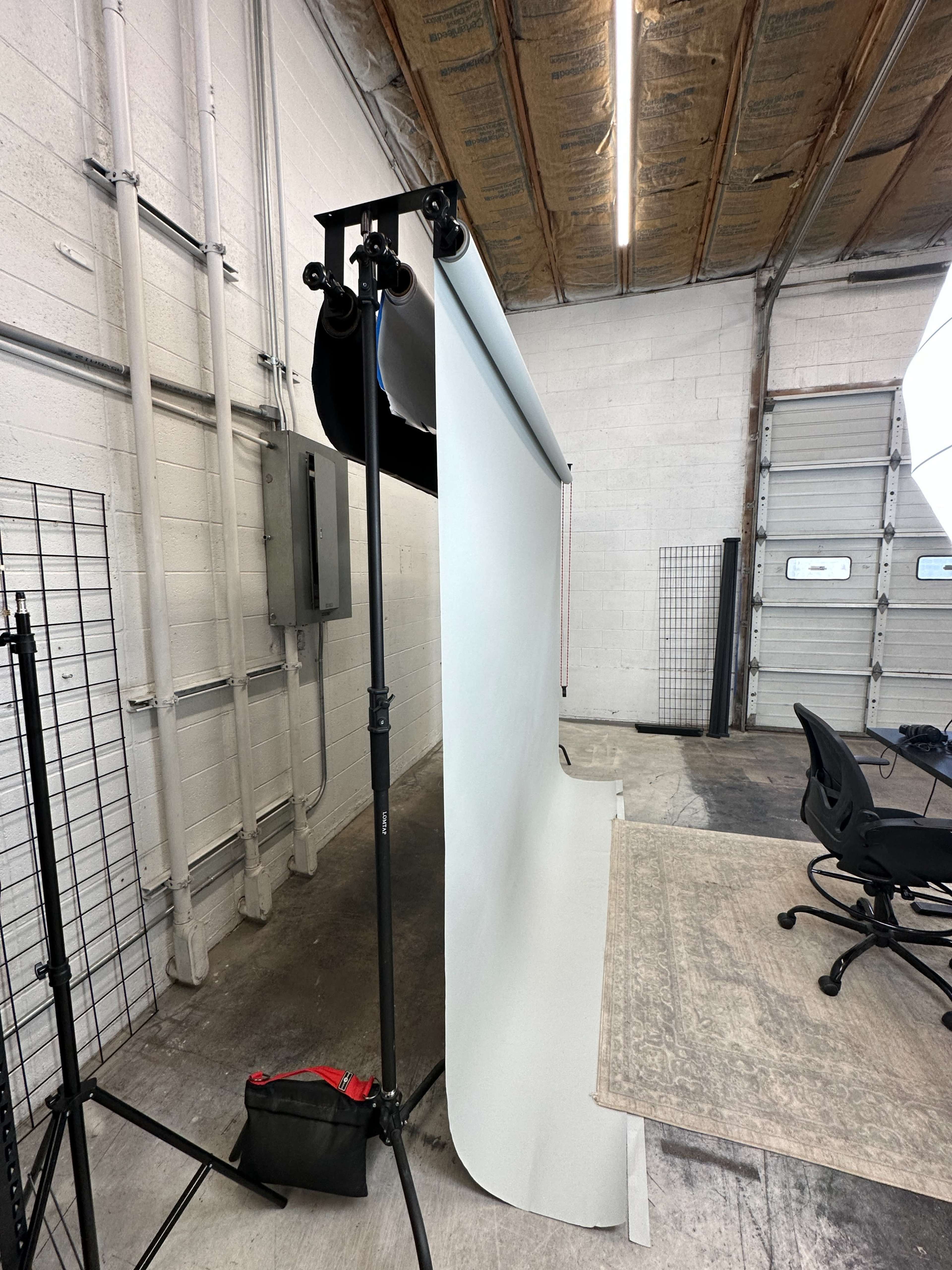 A photography studio features a rolling backdrop stand with a white background and empty chairs arranged nearby.