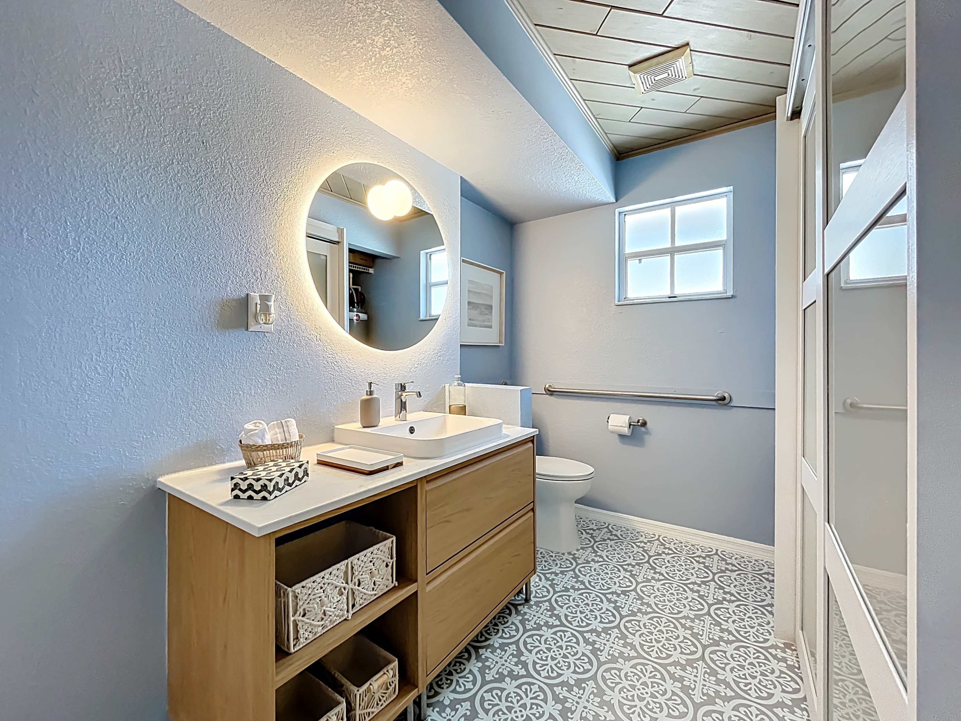 The image shows a modern bathroom featuring a circular mirror, a white sink with a wooden vanity, and patterned tile flooring.