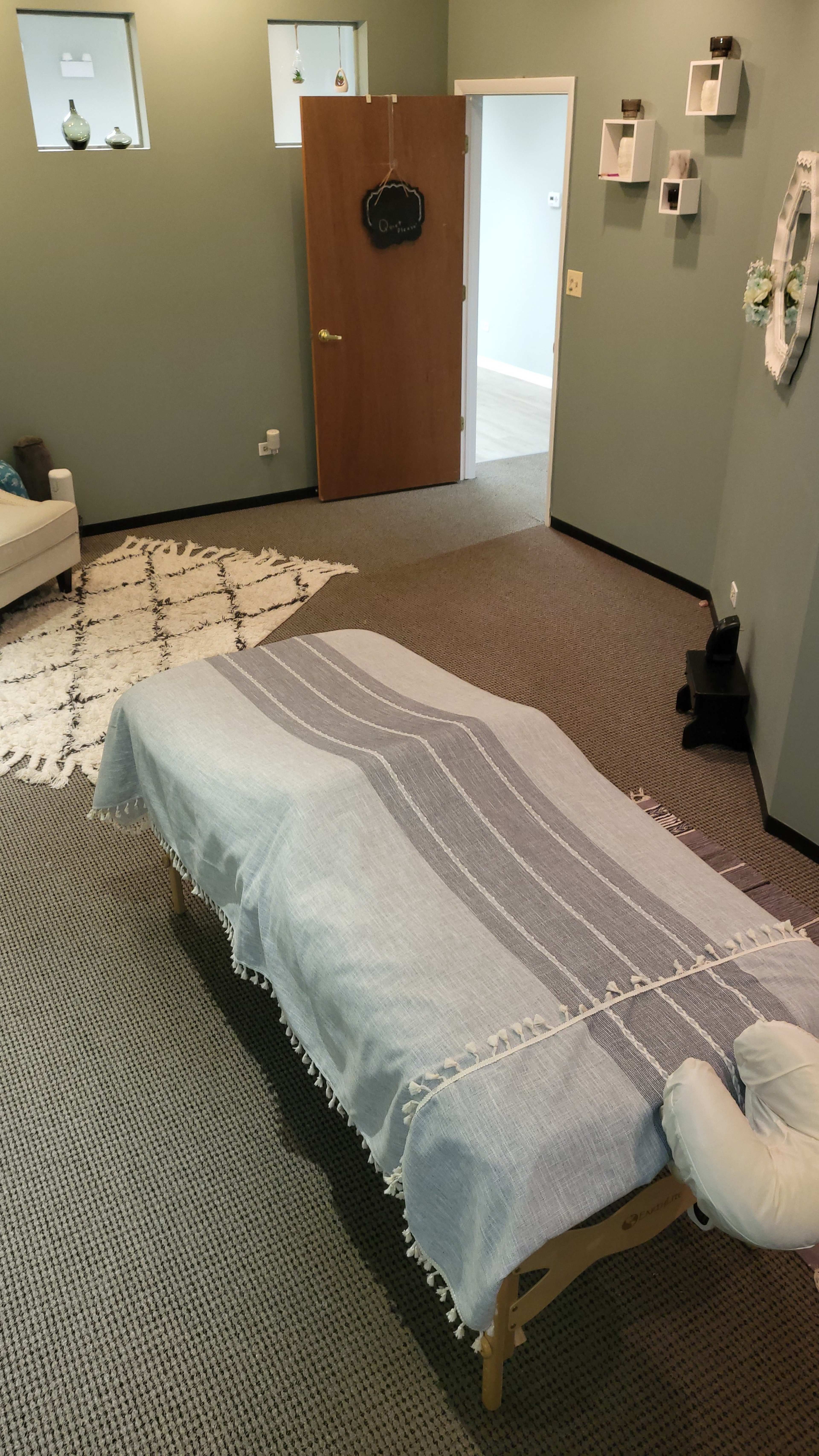 A massage therapy room features a treatment table covered with a light blue blanket, surrounded by neutral-colored walls and simple decor, including a door and a small seating area.