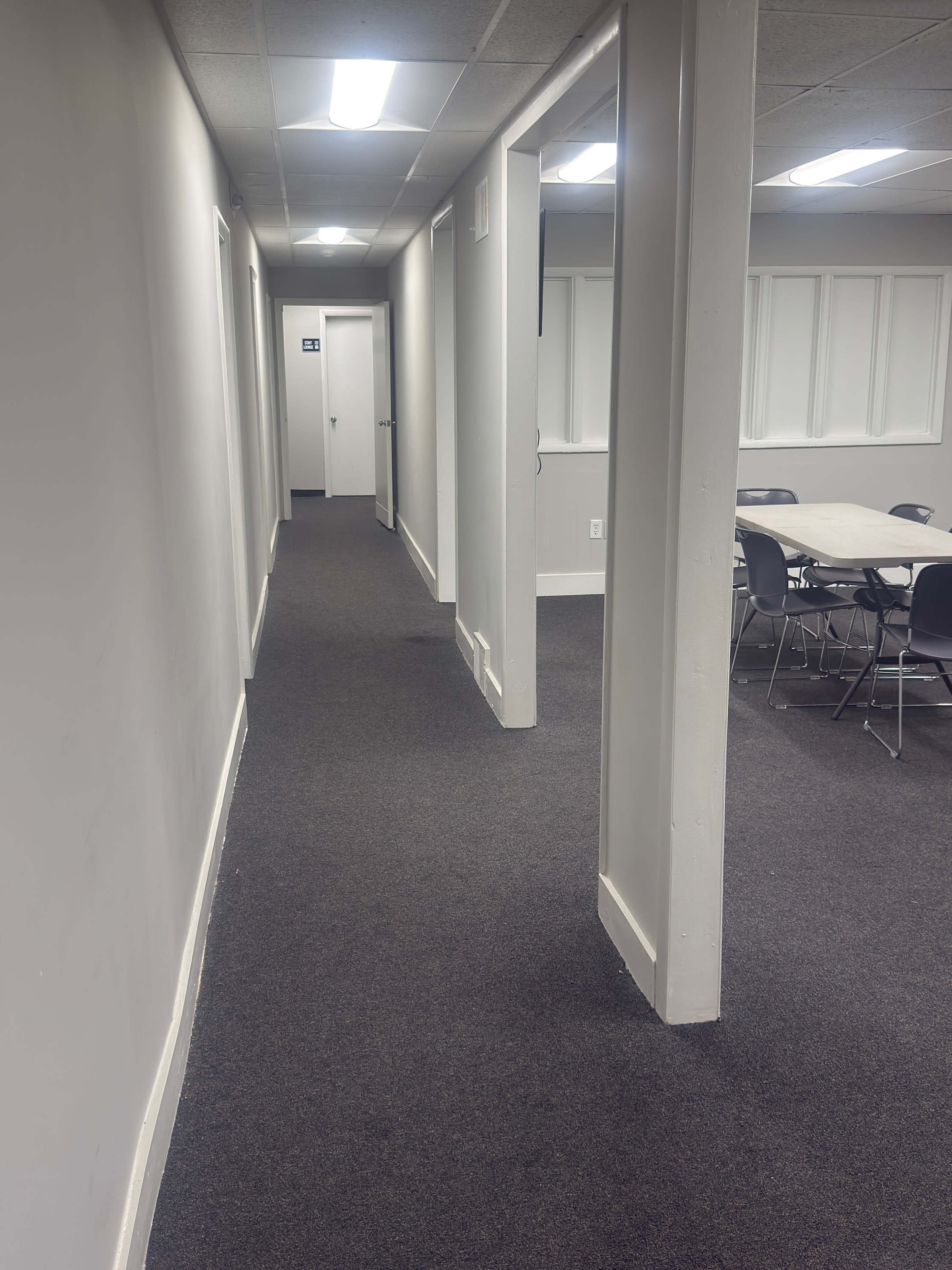 The image shows a long, narrow hallway with gray walls, carpeted flooring, a door at the end, and a meeting room with a table and chairs on the right side.