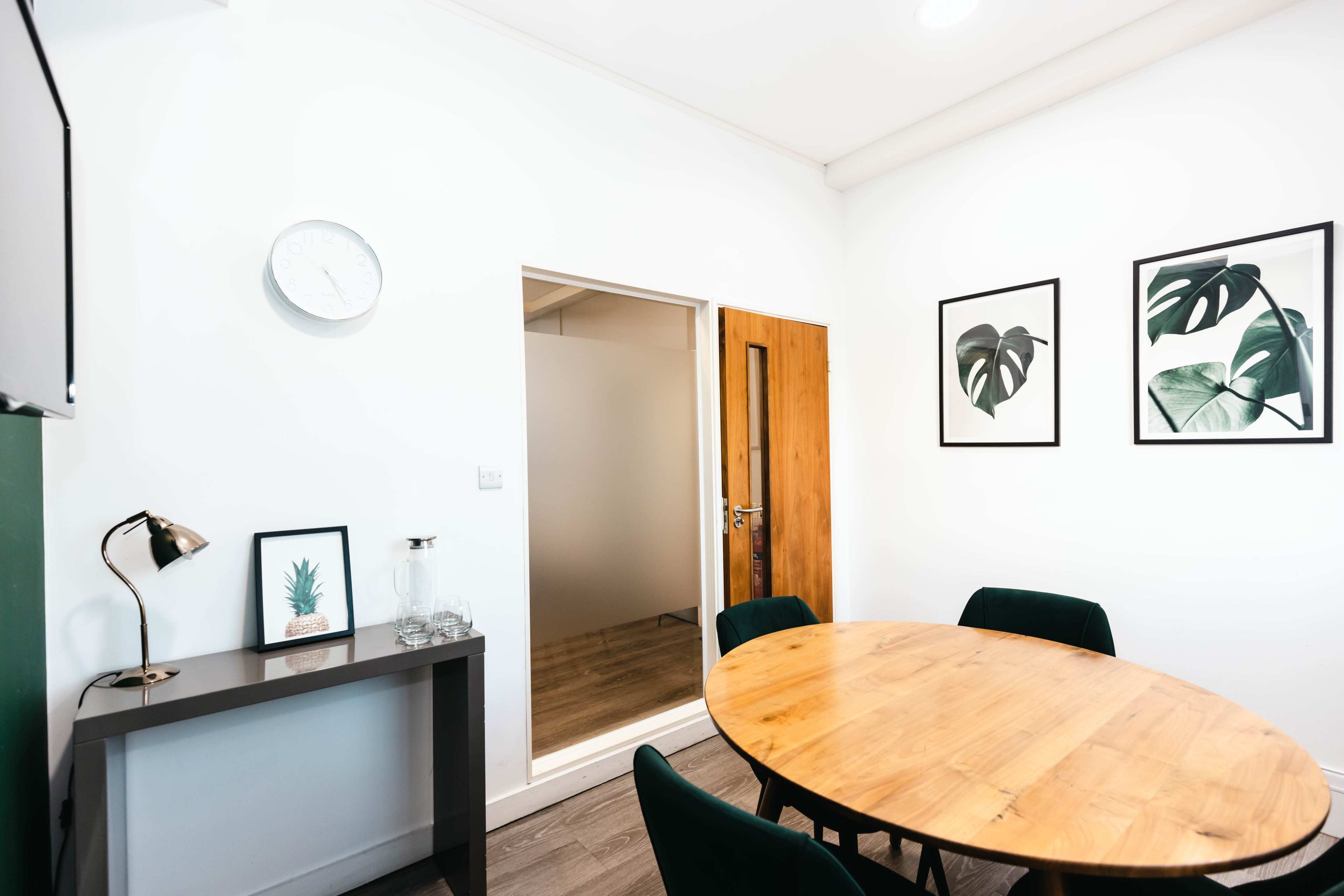 A round wooden table is surrounded by dark green chairs in a room with a clock, a framed plant print, and a door leading to another space.