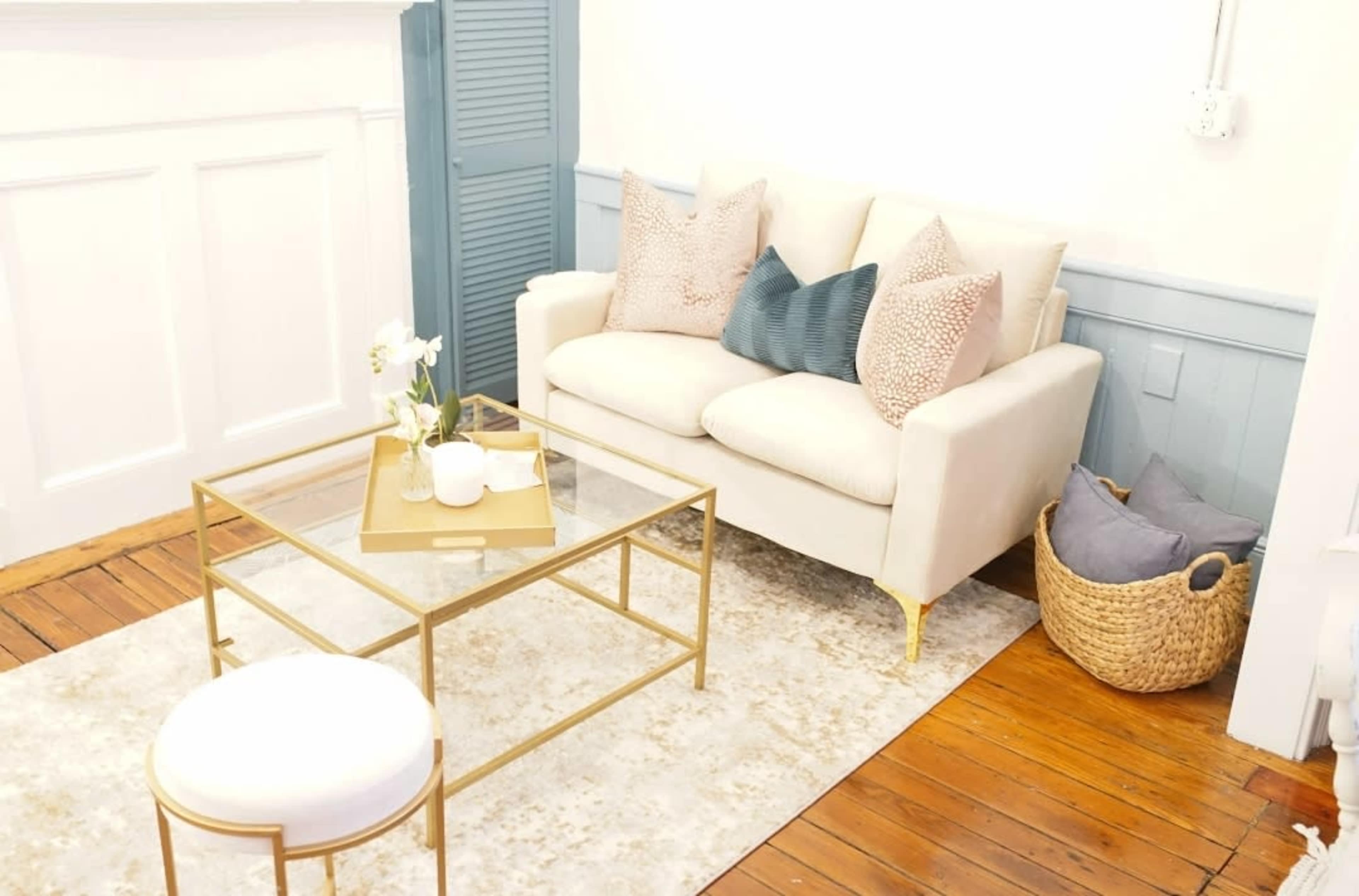 A light-colored sofa with decorative pillows sits across from a glass-top coffee table in a room with wooden flooring and a woven basket.